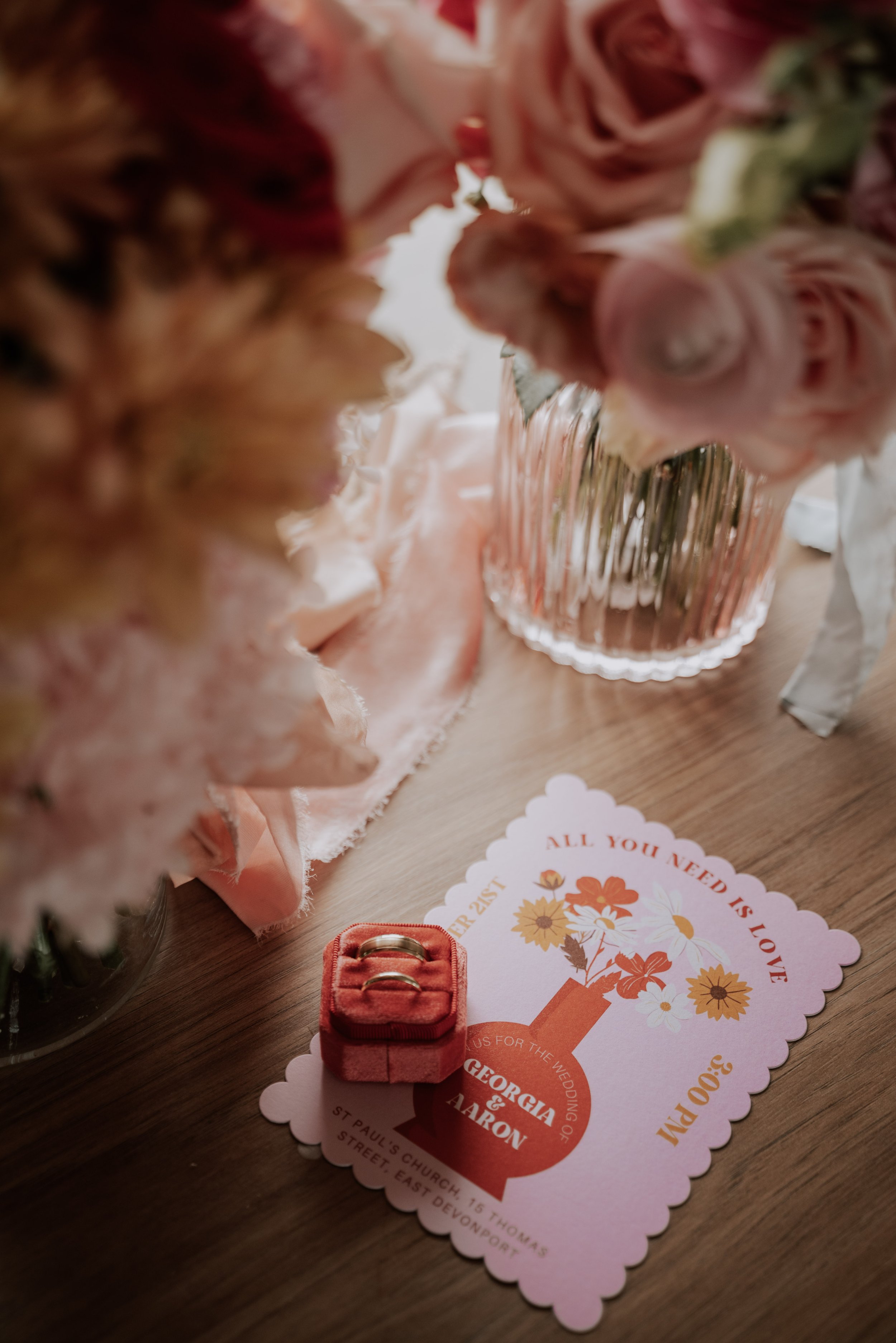 Wedding bouquets and pink wedding stationary sit on a bench with the wedding rings in a red velvet box