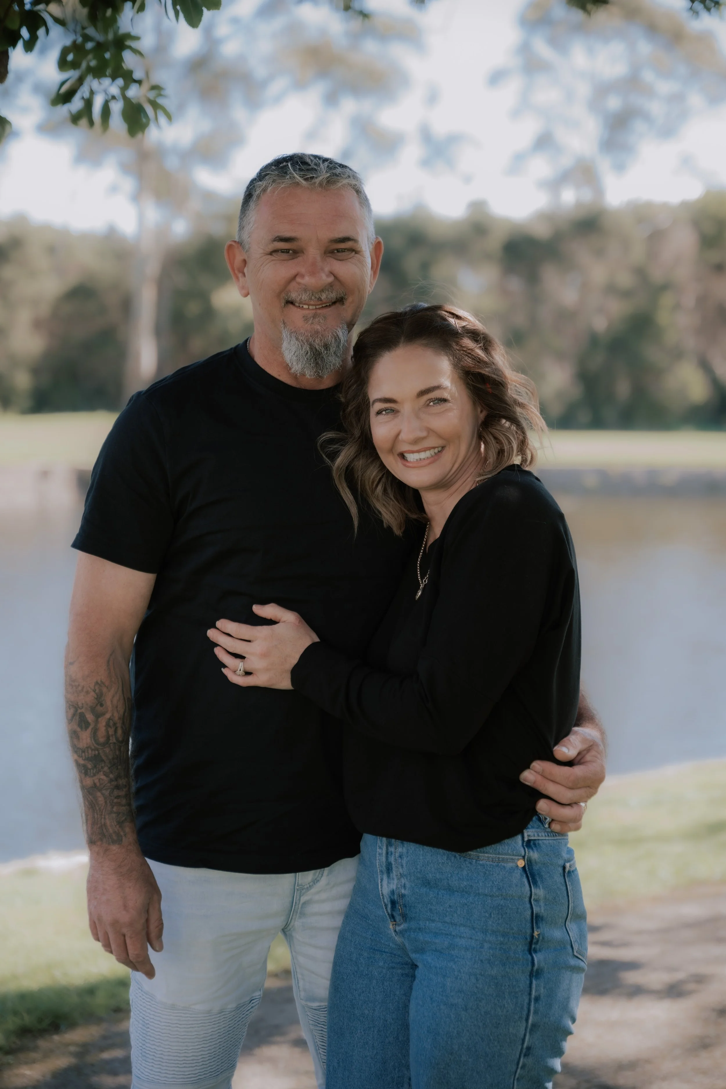 A middle-aged man and woman smiling and embracing outdoors near a body of water with trees in the background.