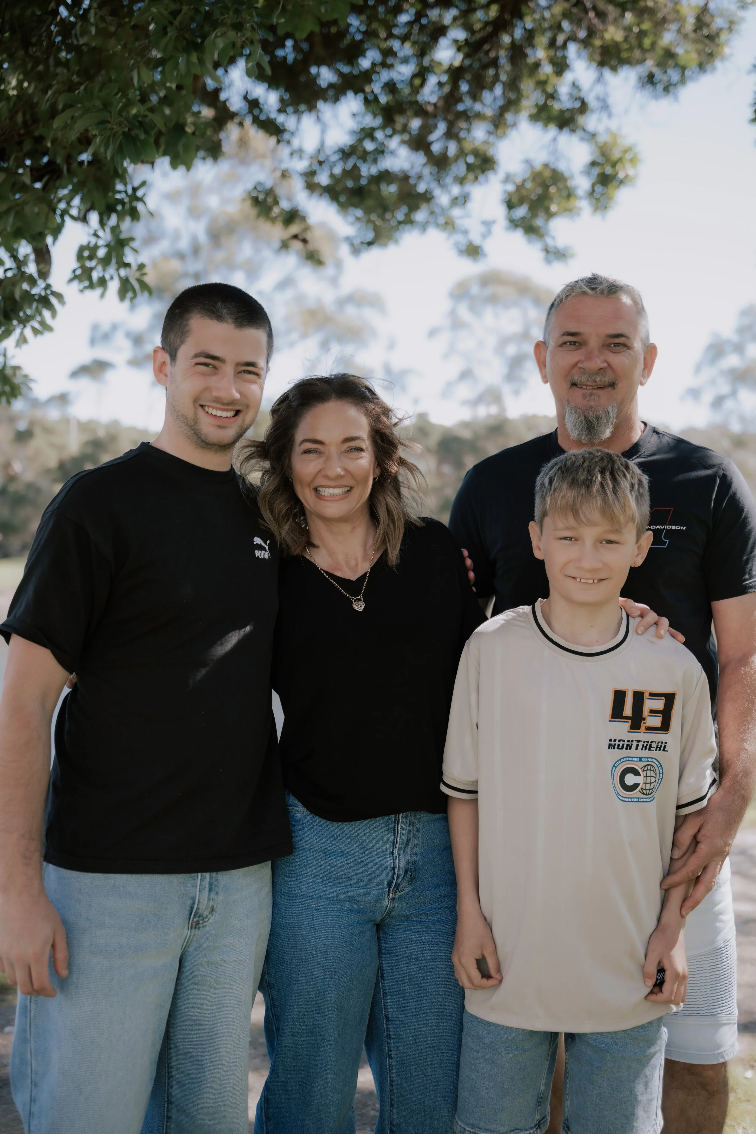 A diverse family standing outdoors under a tree, smiling at the camera. The group includes a young man, a woman, an older man, and a young boy, all dressed casually.