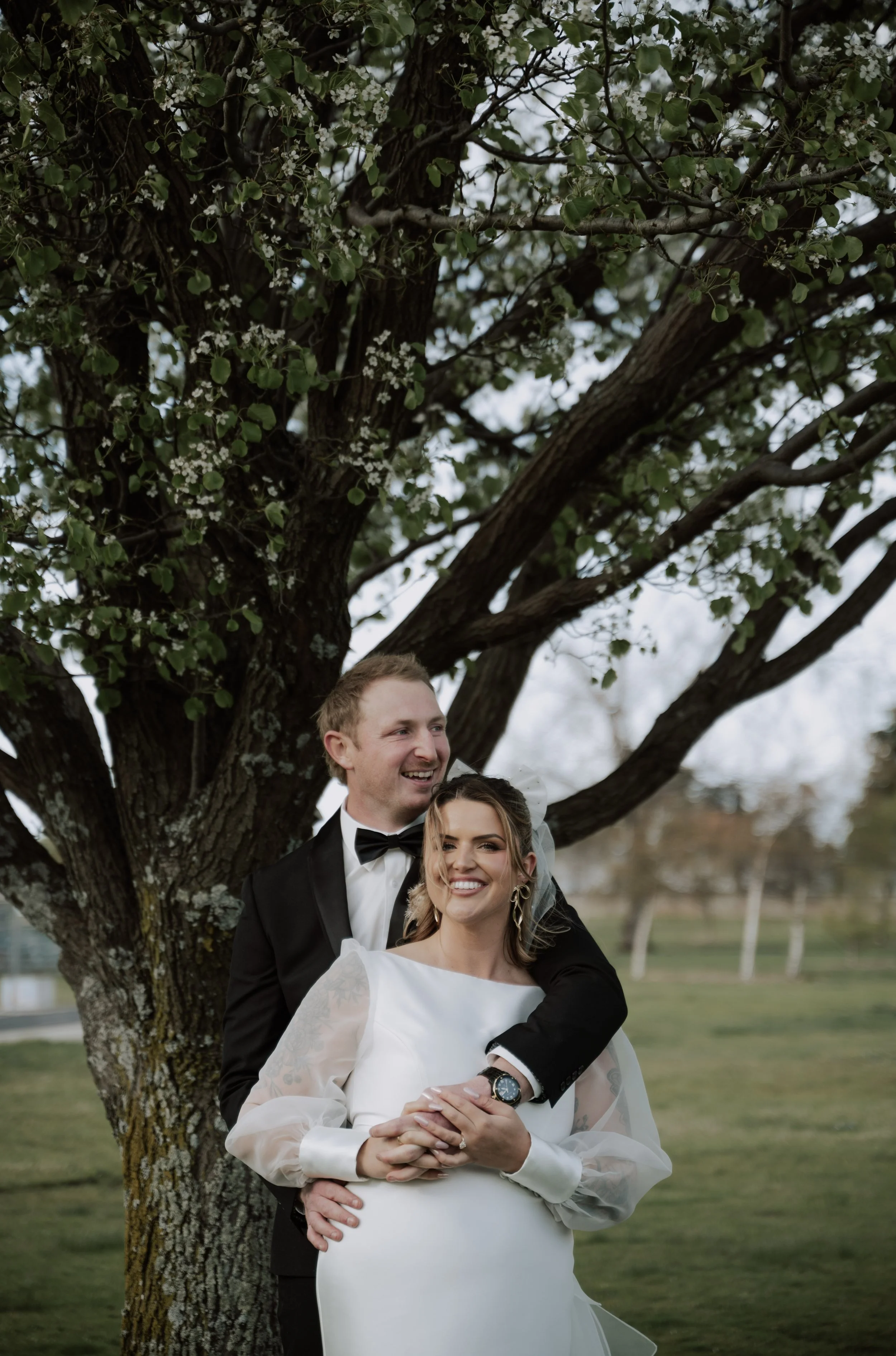 A bride and groom face the camera while hugging candidly and smiling beneath a tall green tree in a park