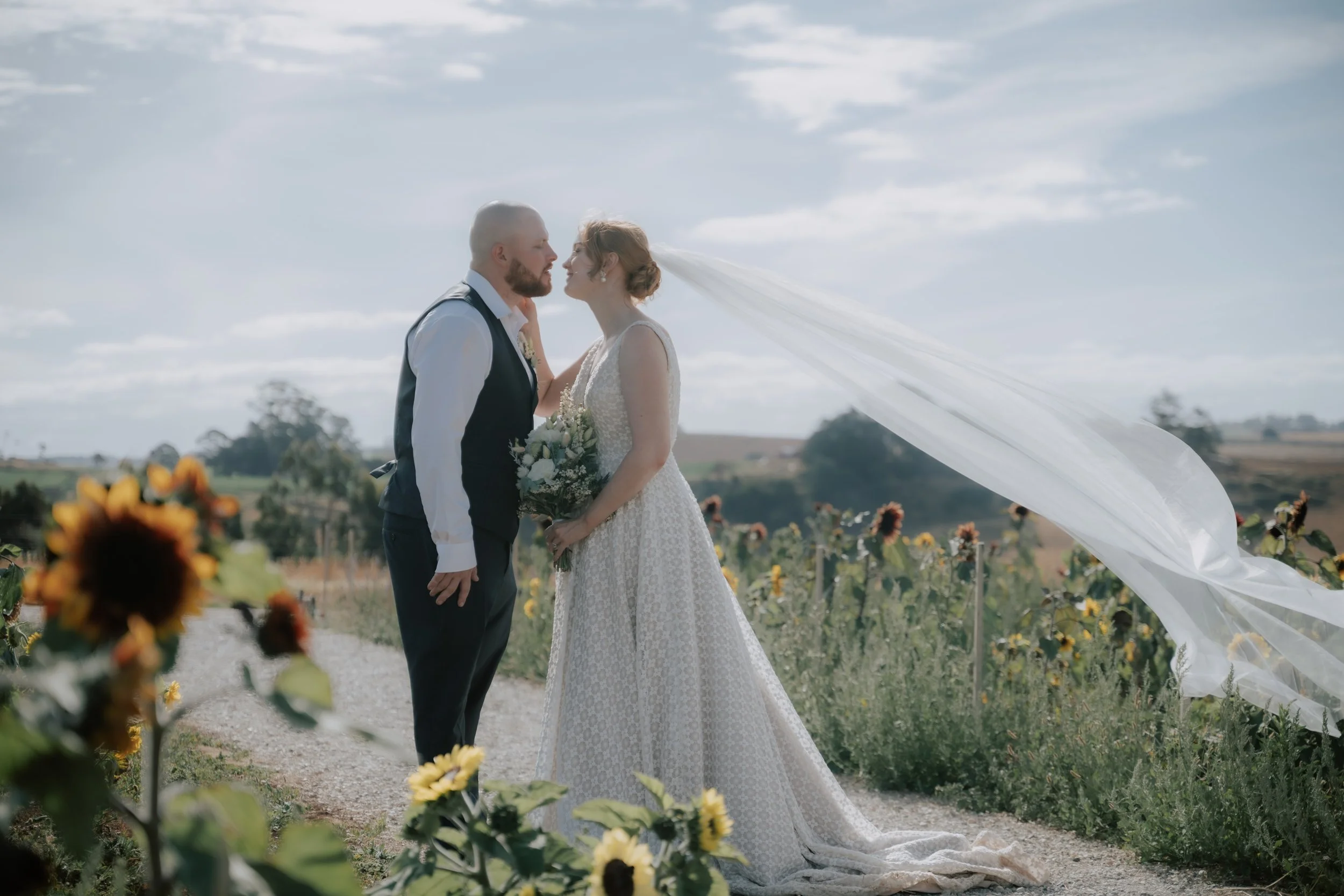 A bride and groom standing close together outdoors in a field of sunflowers, facing each other with foreheads nearly touching. The bride is wearing a white lace wedding dress and veil, holding a bouquet, and the groom is dressed in a white shirt with