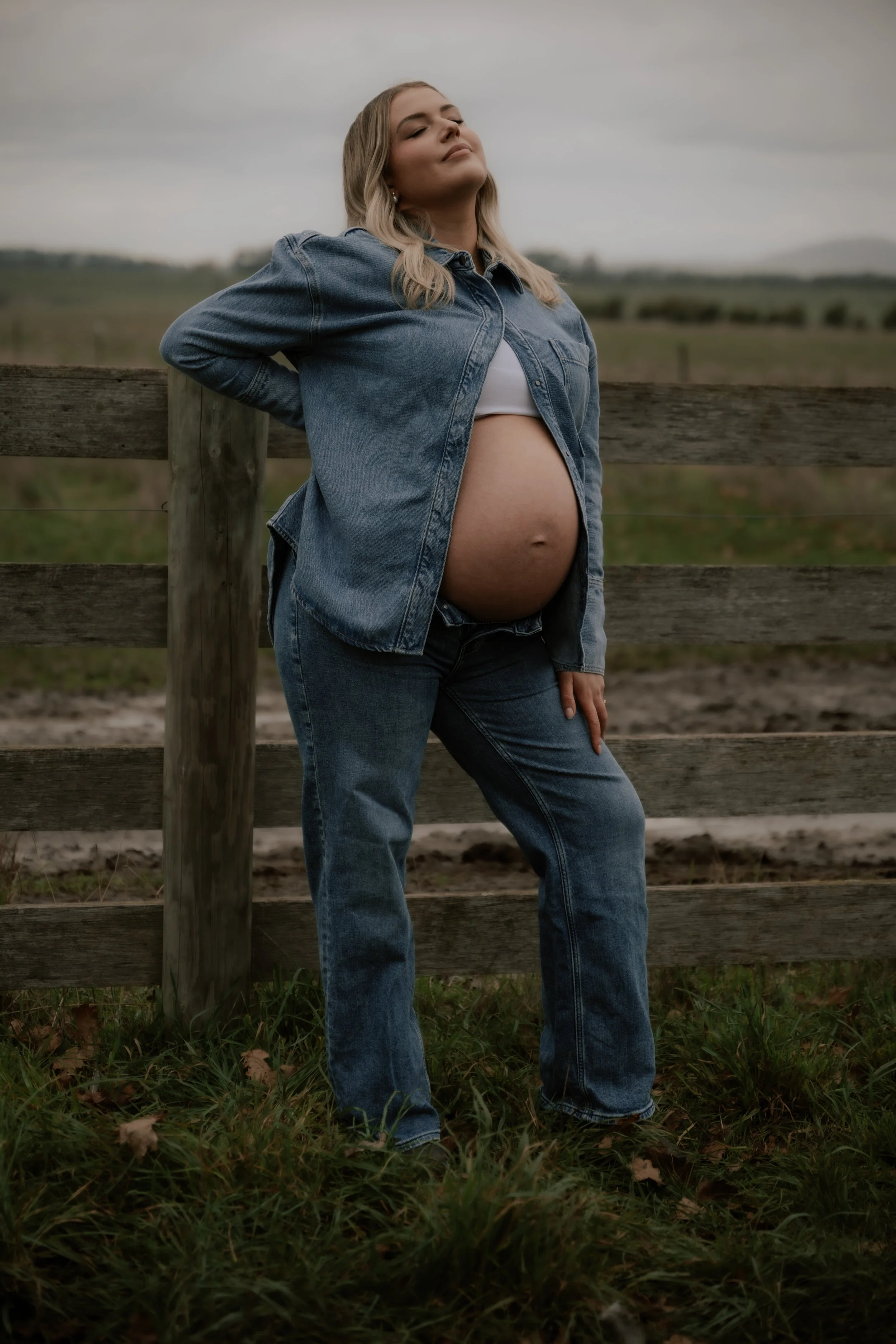 A pregnant woman with blonde hair stands outdoors near a wooden fence, with her eyes closed and a content expression, wearing a denim jacket and jeans, over a white top, in a rural setting with green grassy field and cloudy sky.
