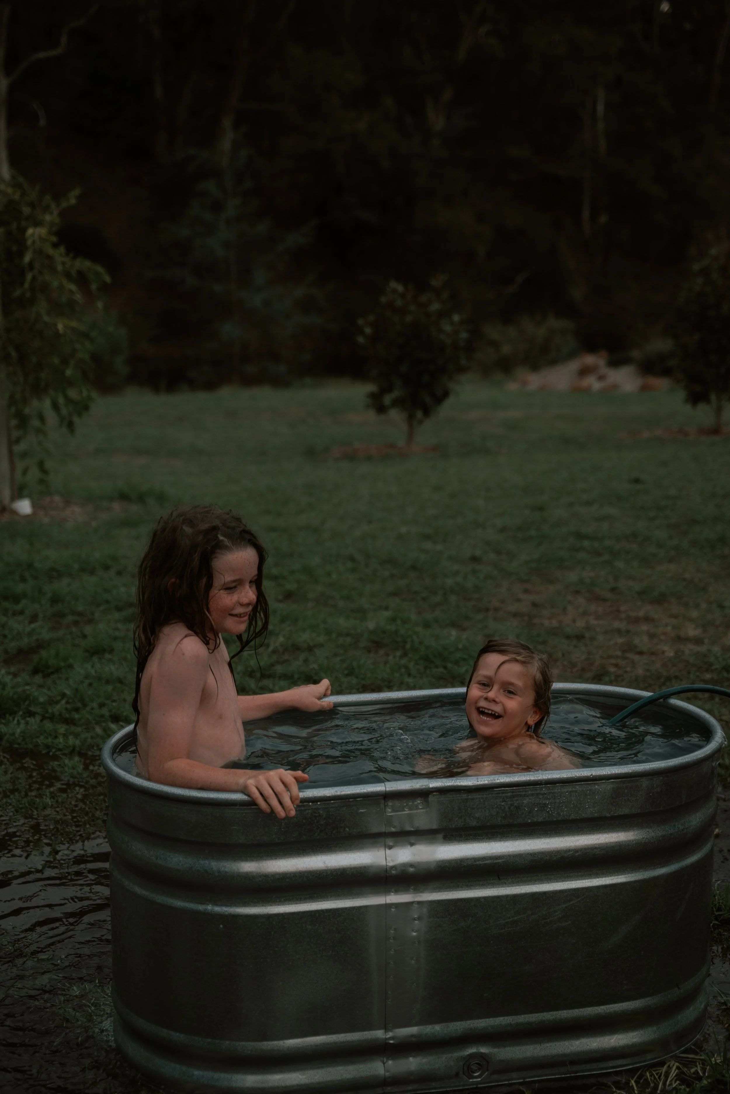 Two children with wet hair enjoying a bath in a metal tub outdoors, surrounded by grass and trees during dusk or evening.