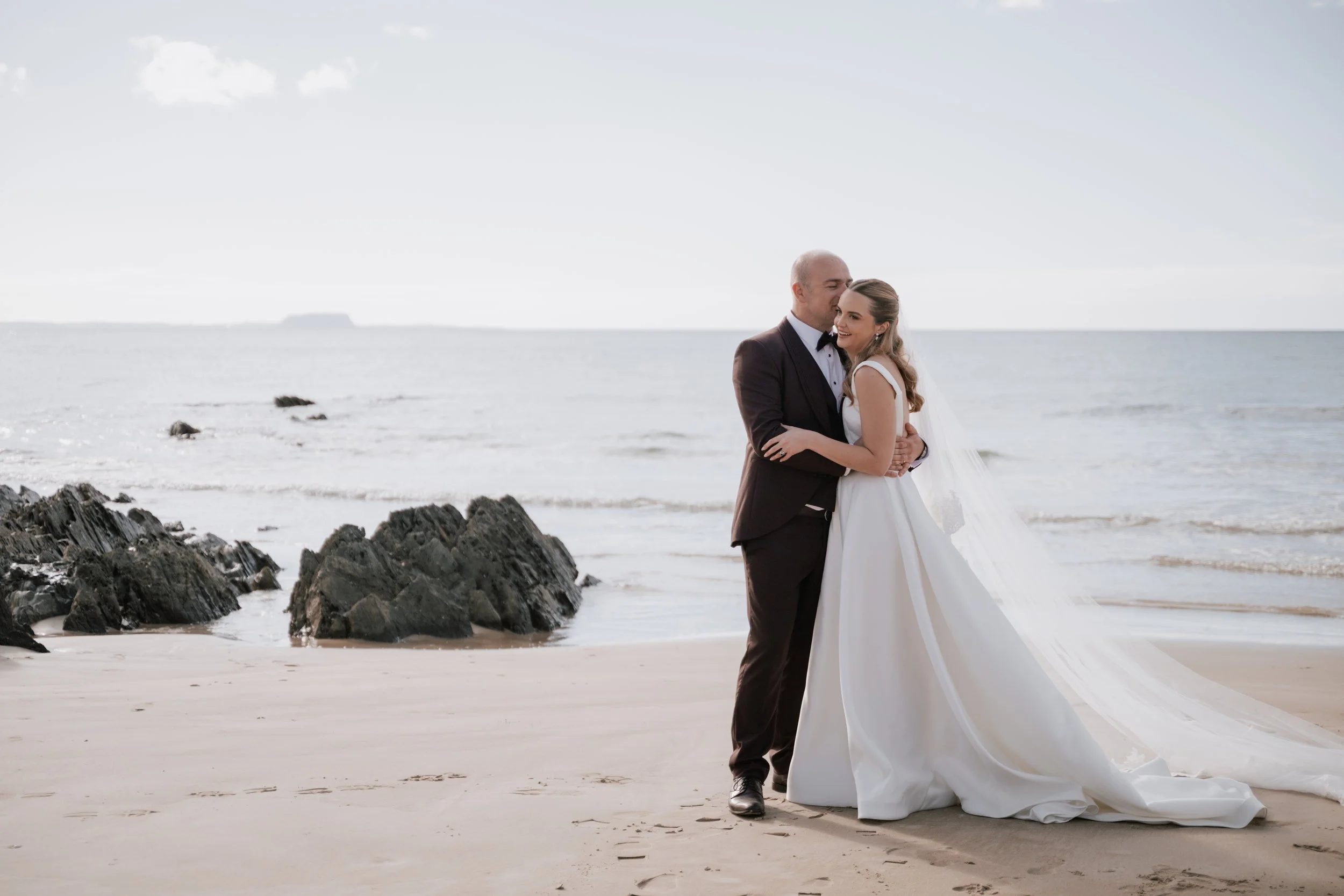 A bride and groom hugging on a beach during their wedding, with rocks in the foreground and the ocean in the background.