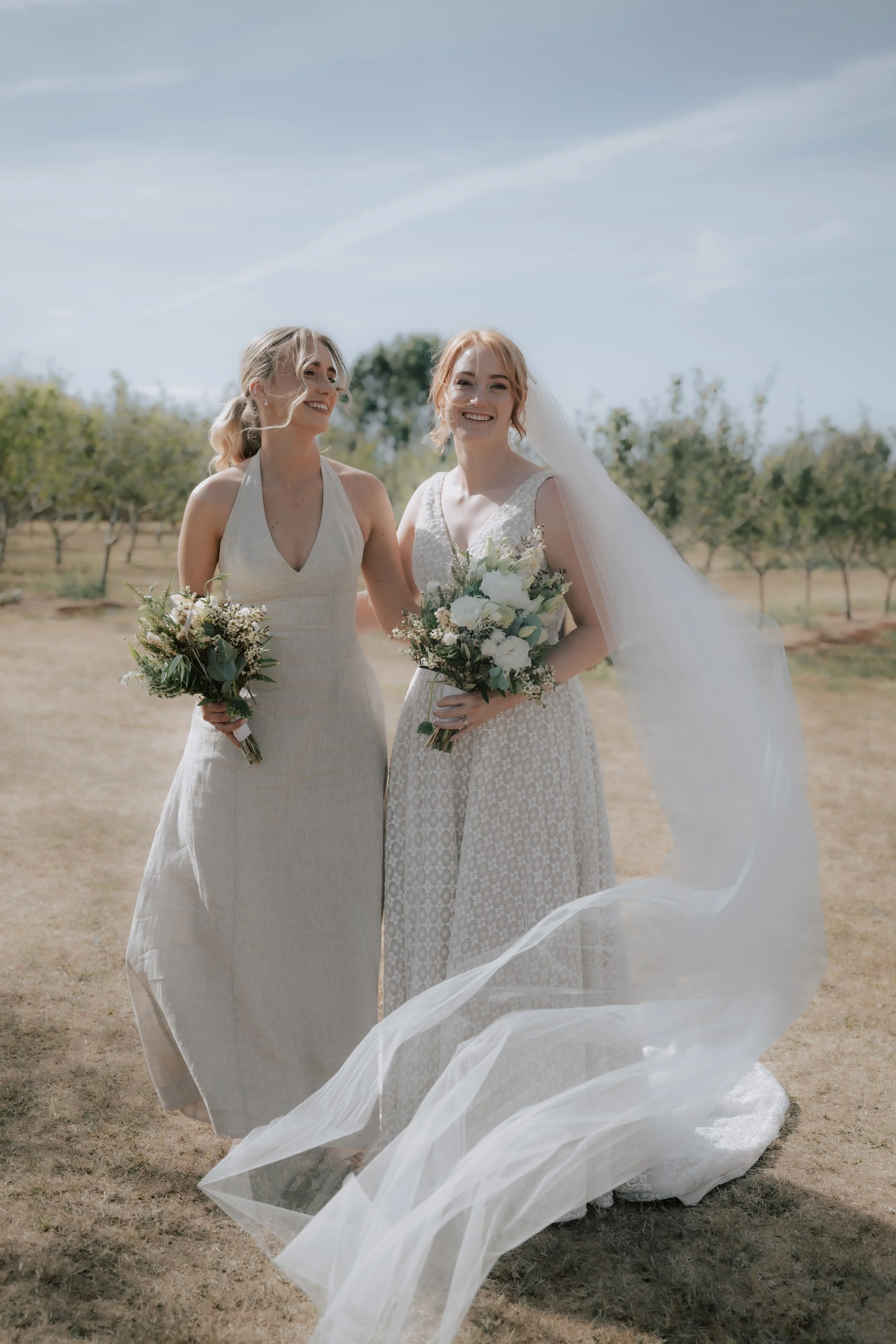 Two women in wedding dresses standing outdoors in a field with trees in the background. They are smiling and holding bouquets of flowers, with wedding veil flowing in the breeze.