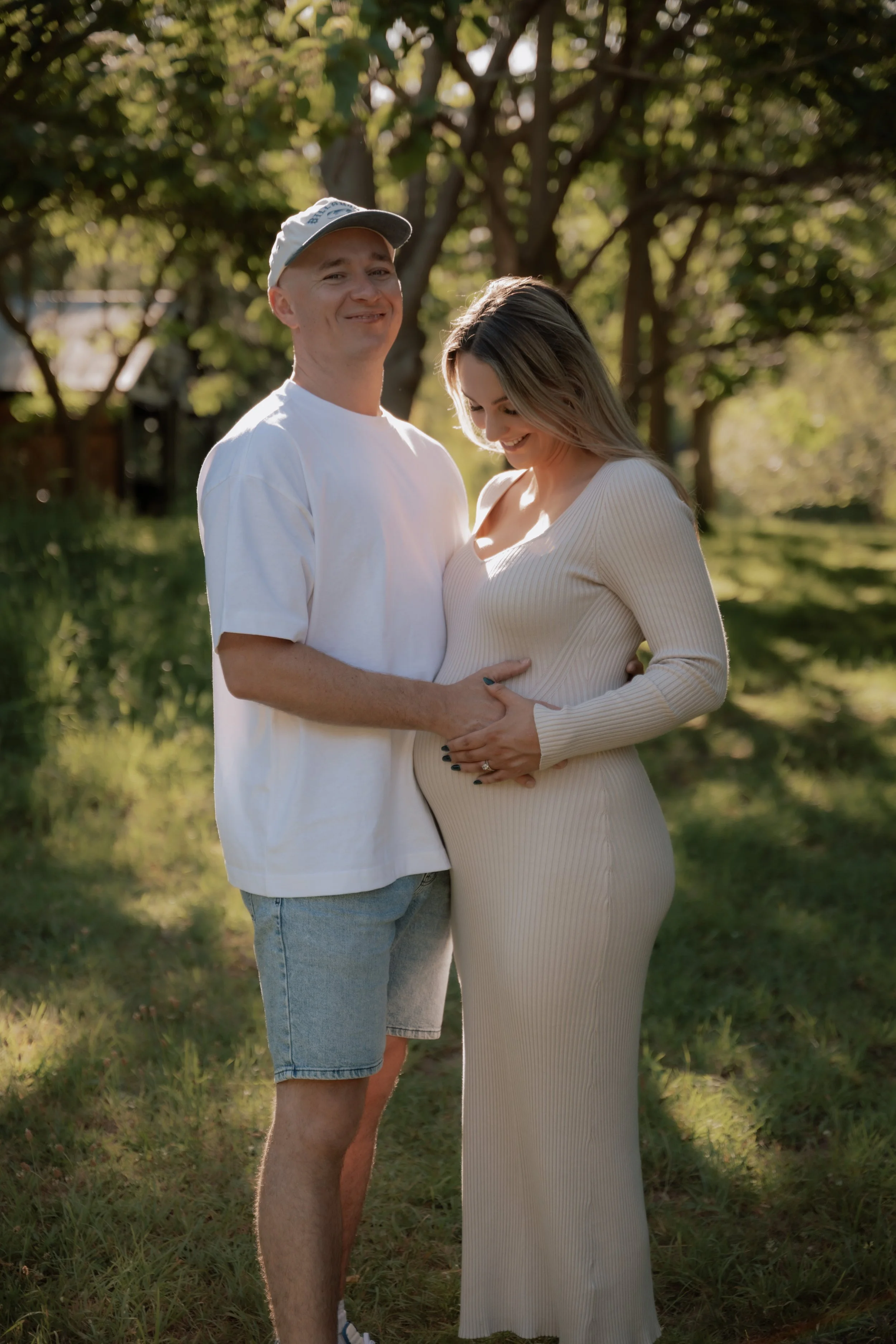 A smiling man wearing a white T-shirt, denim shorts, and a cap, standing next to a pregnant woman in a beige dress. They are outdoors in a park with trees and sunlight filtering through the leaves.