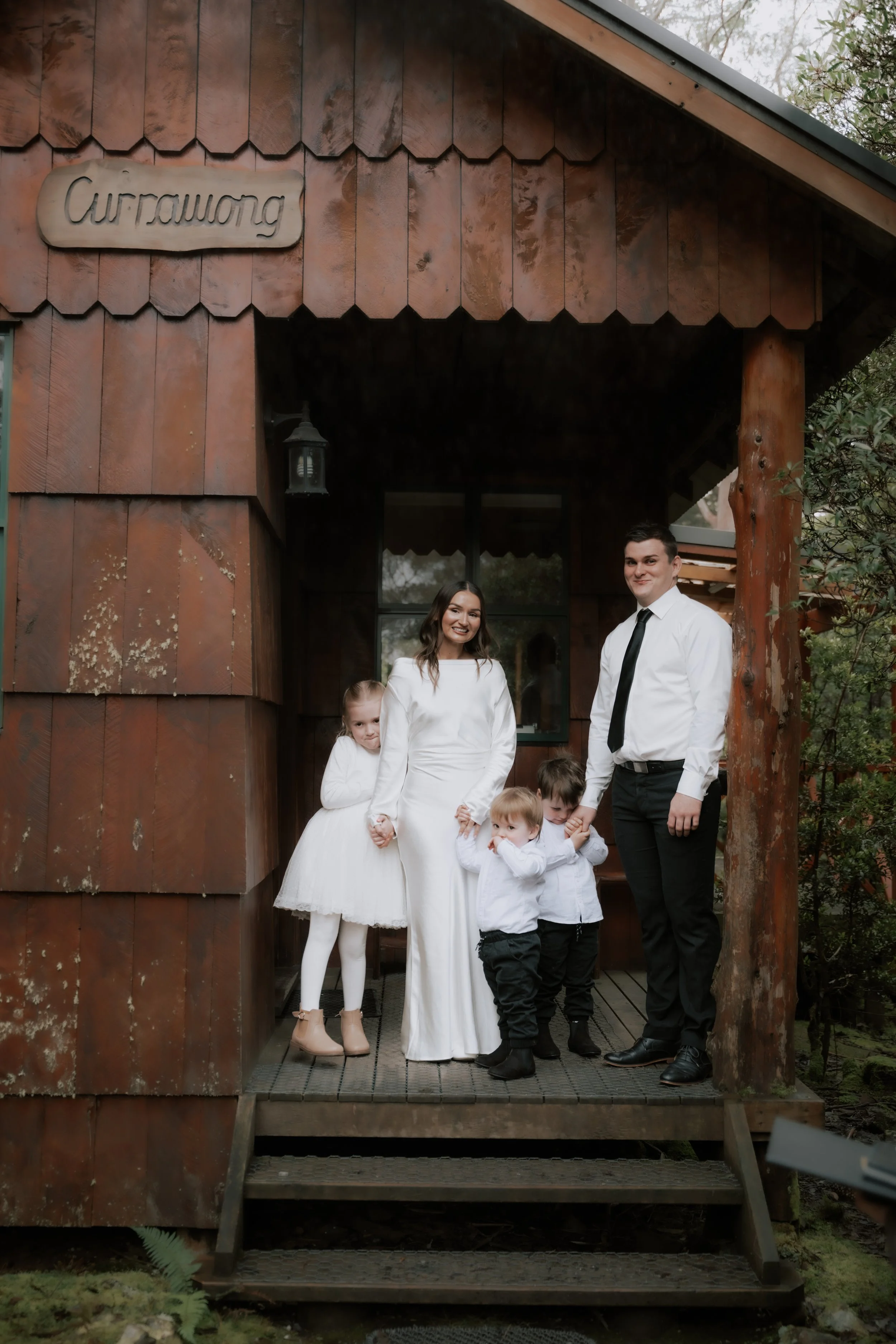 A family of six standing on the steps of a wooden house, with the women and children dressed in white, and the man in a white shirt and black tie, posing for a photo outdoors in front of a rustic wooden building.