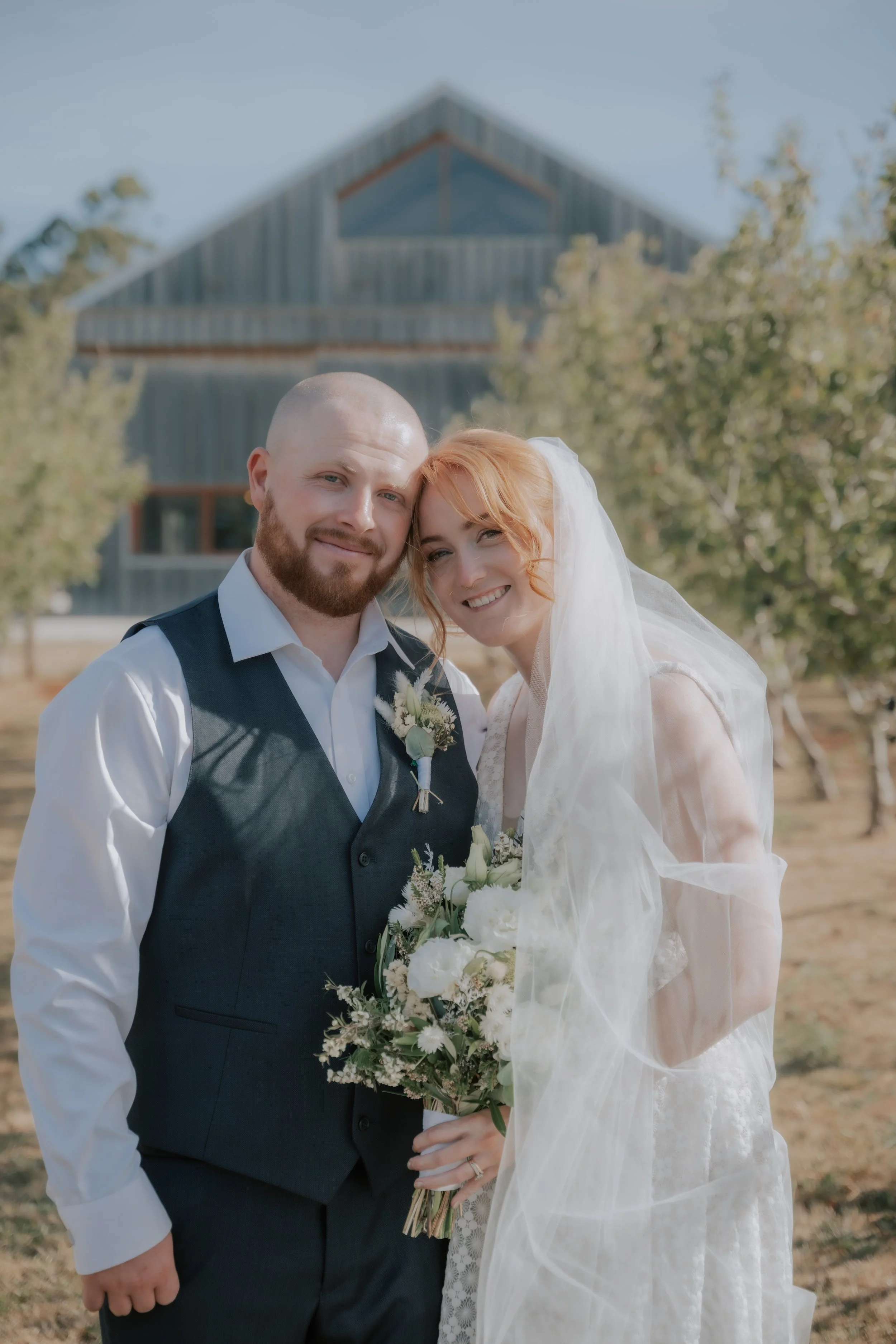 A bride and groom posing outdoors with a barn in the background, smiling and holding a bouquet of white flowers.