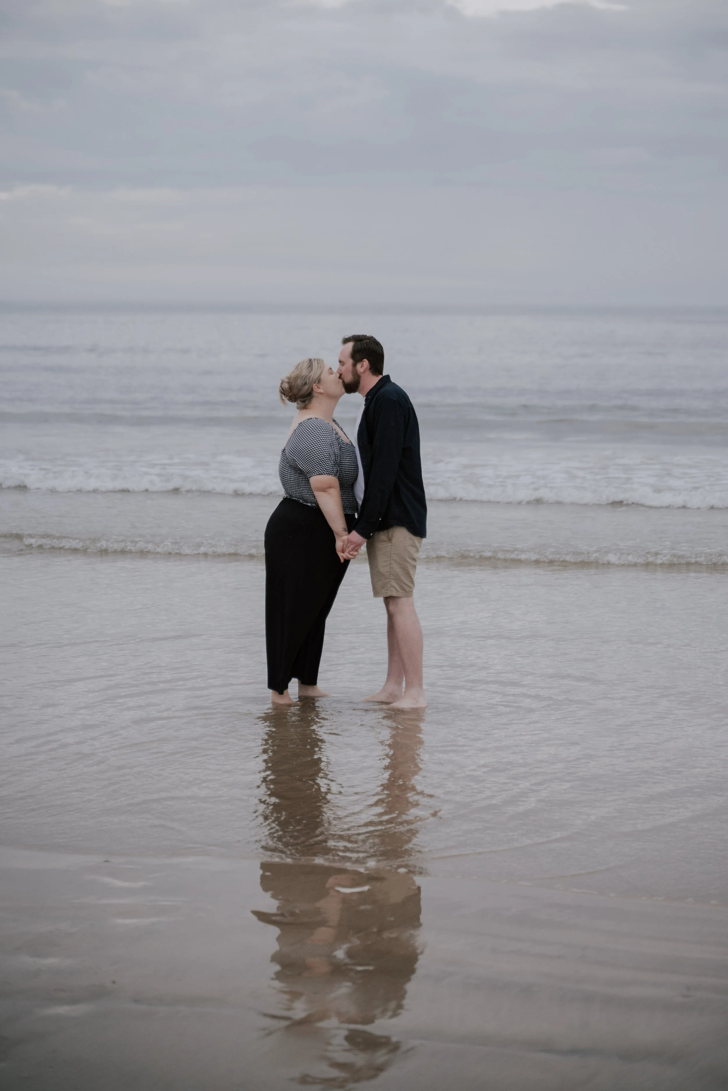 A couple stand in the shallow ocean and kiss to celebrate their engagement, on a cloudy day