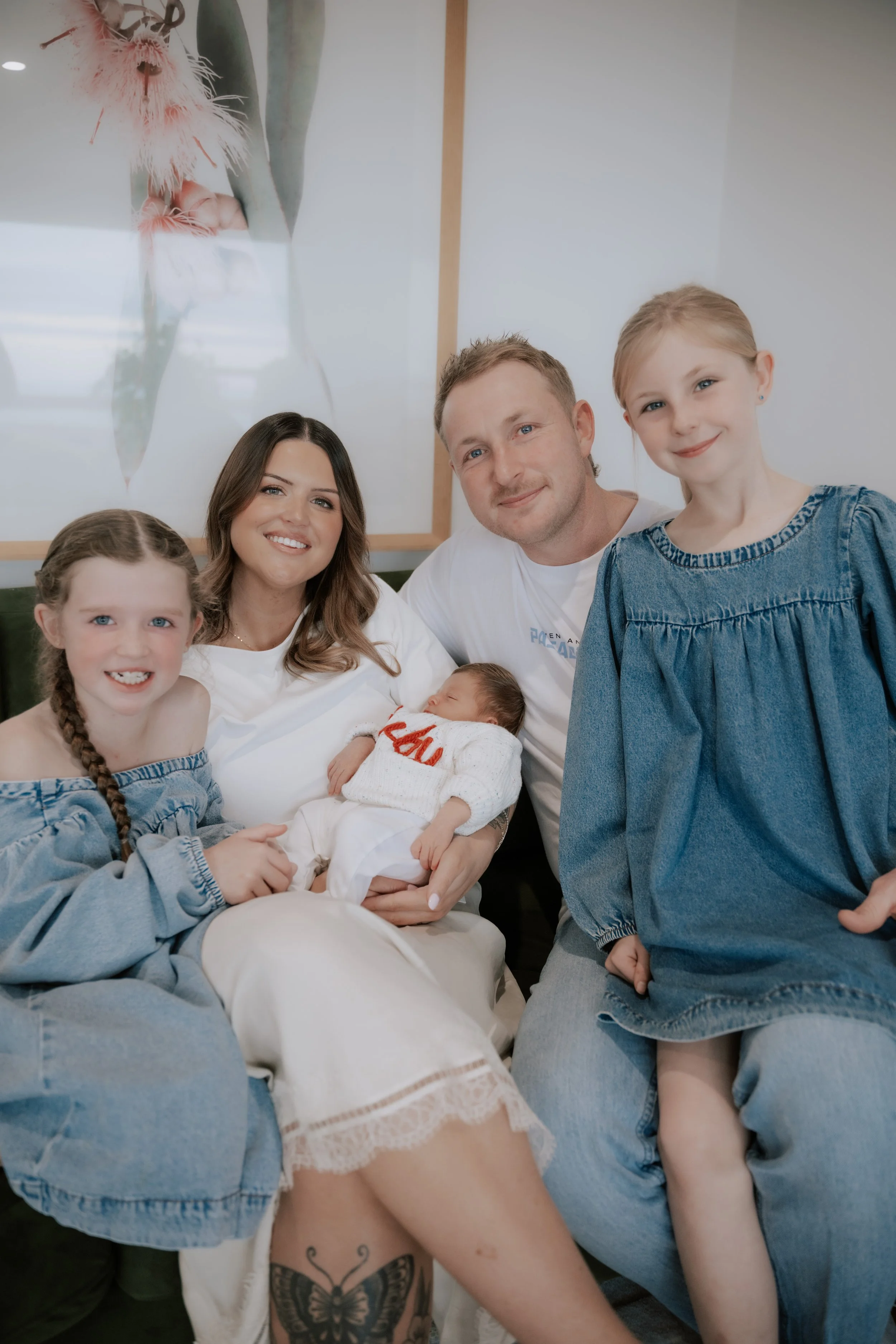 A family of five poses together in a room with a large botanical poster on the wall. They include an adult woman, an adult man, two young girls, and an infant. The woman is holding the baby and smiling, the man and girl on the right are smiling, and 