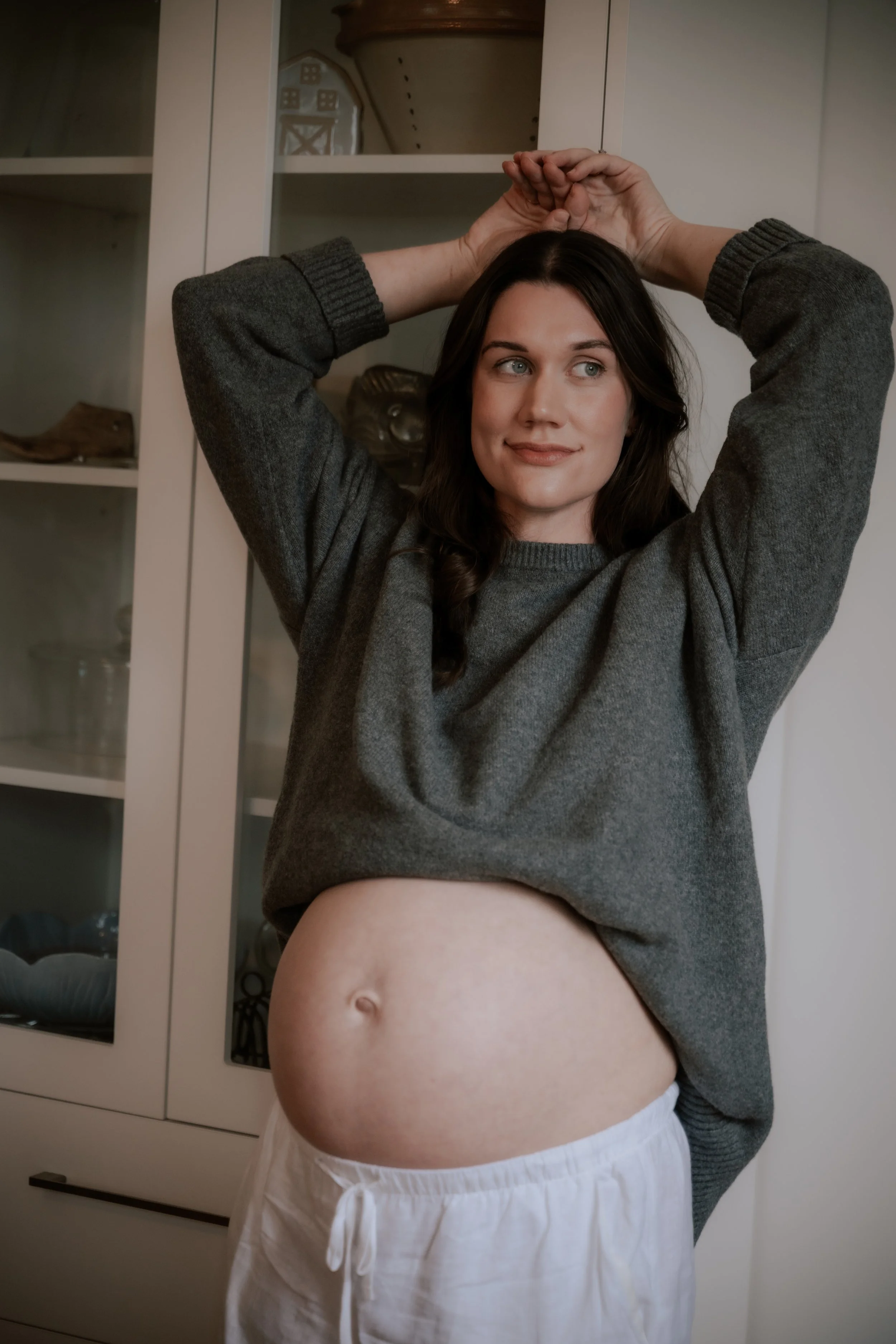 A woman with dark hair wearing a gray sweater, holding her hands above her head, stands in front of a cabinet. She has a slight smile and is revealing her bare stomach and part of her white shorts.