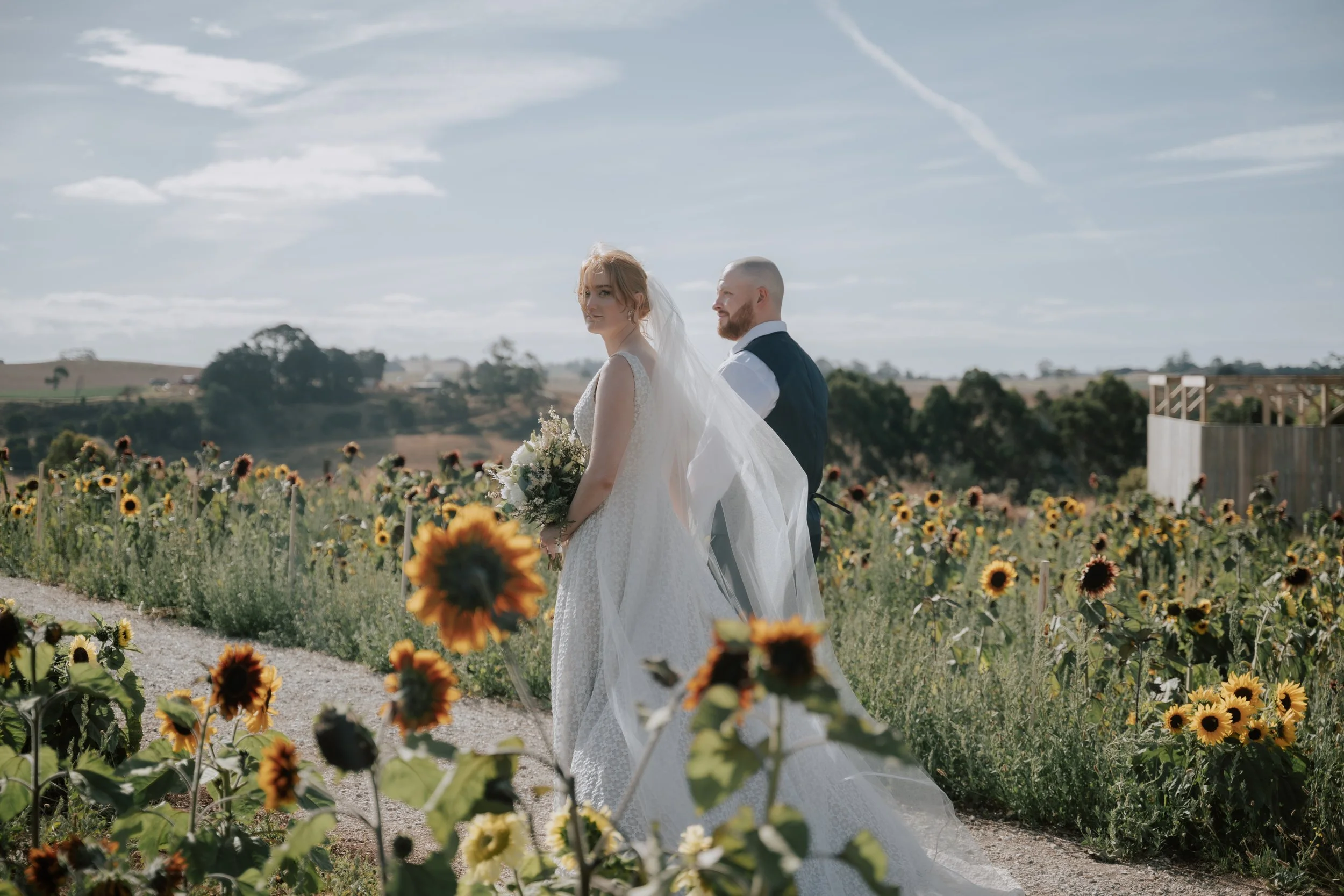 A bride and groom standing in a sunflower field outdoors under a partly cloudy sky, with the bride holding a bouquet of flowers.