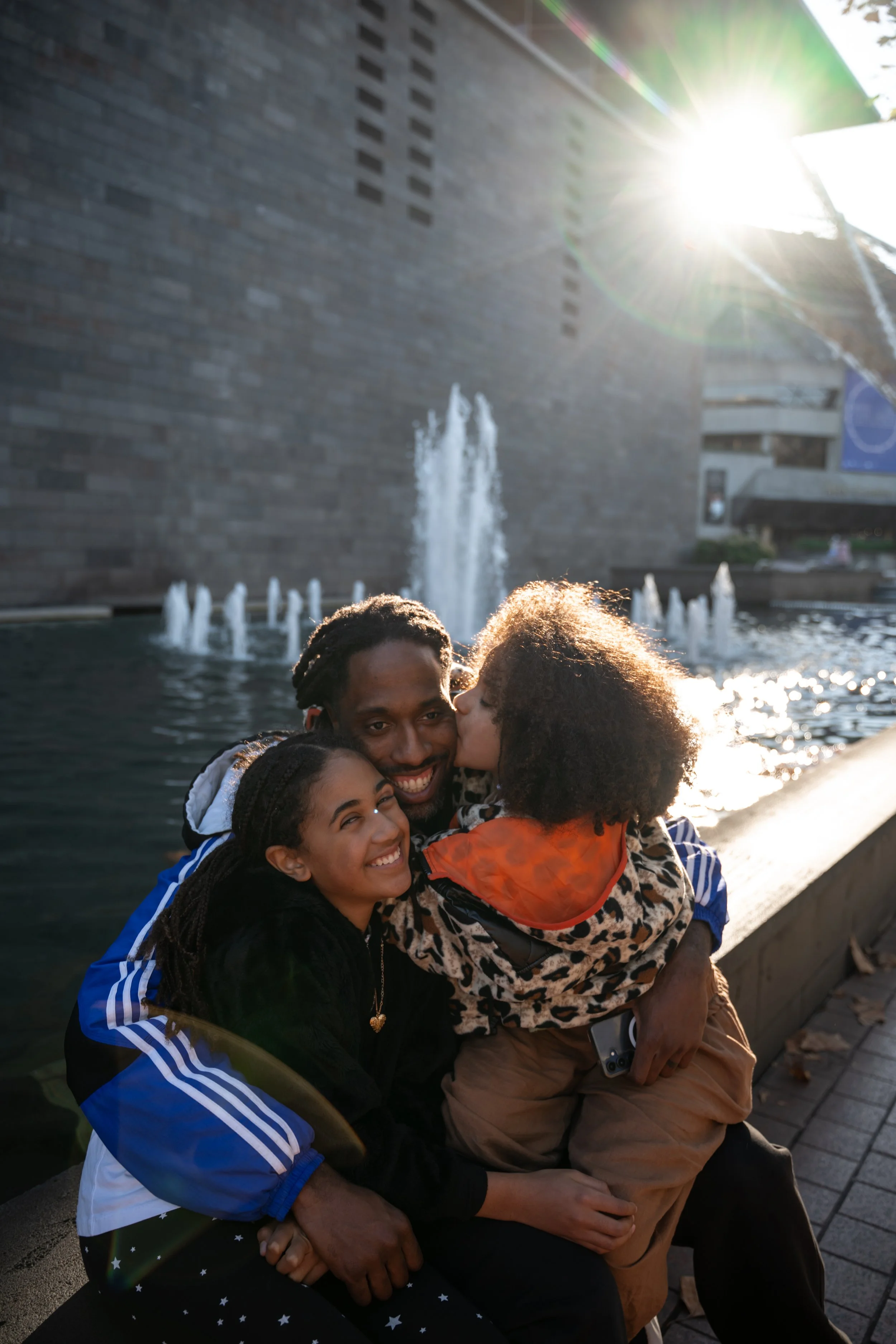 A group of four diverse people hugging and smiling by a fountain with water jets, with the sun shining brightly in the background.