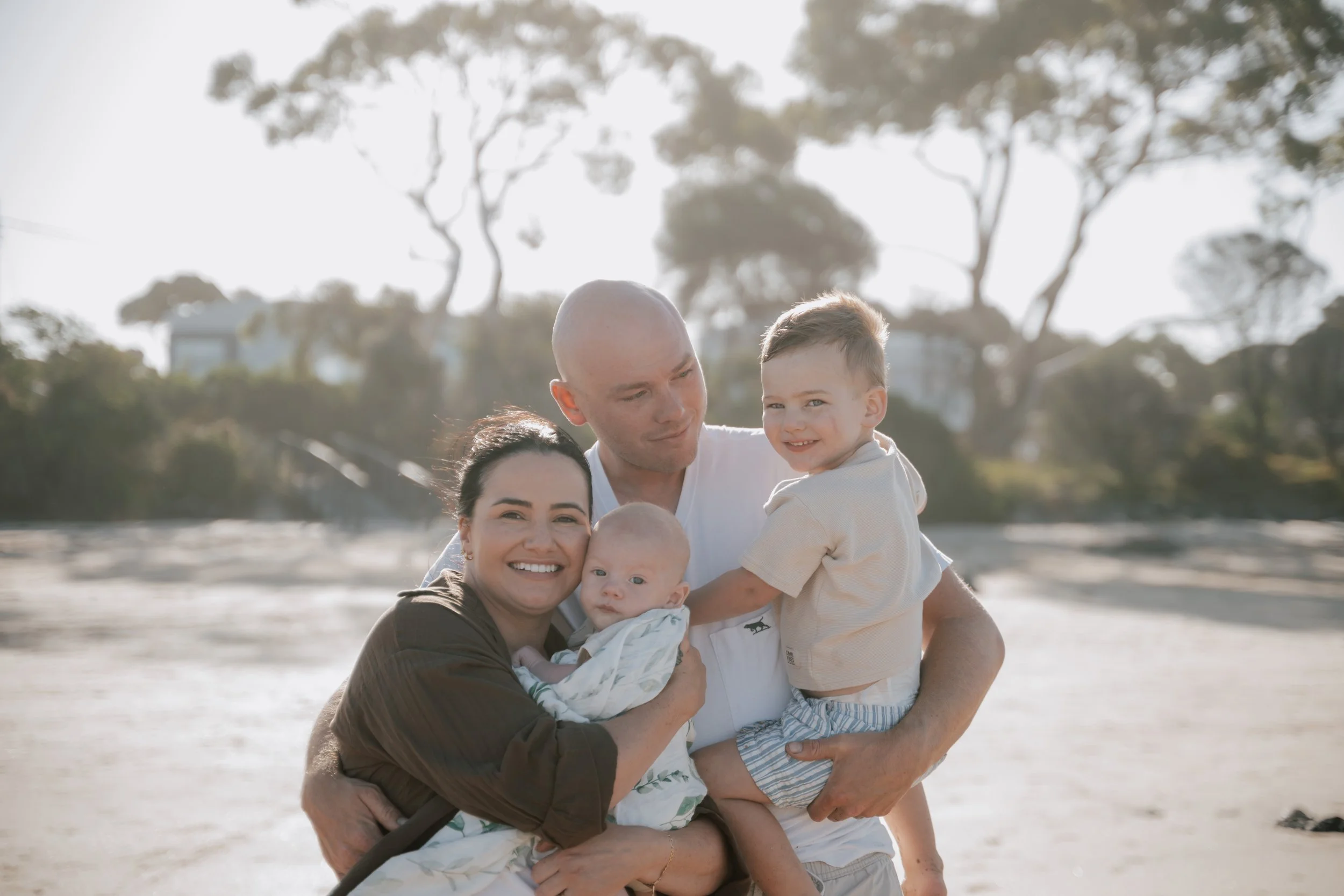 A family of five, including two children and two adults, smiling and hugging outdoors on a beach with trees in the background.