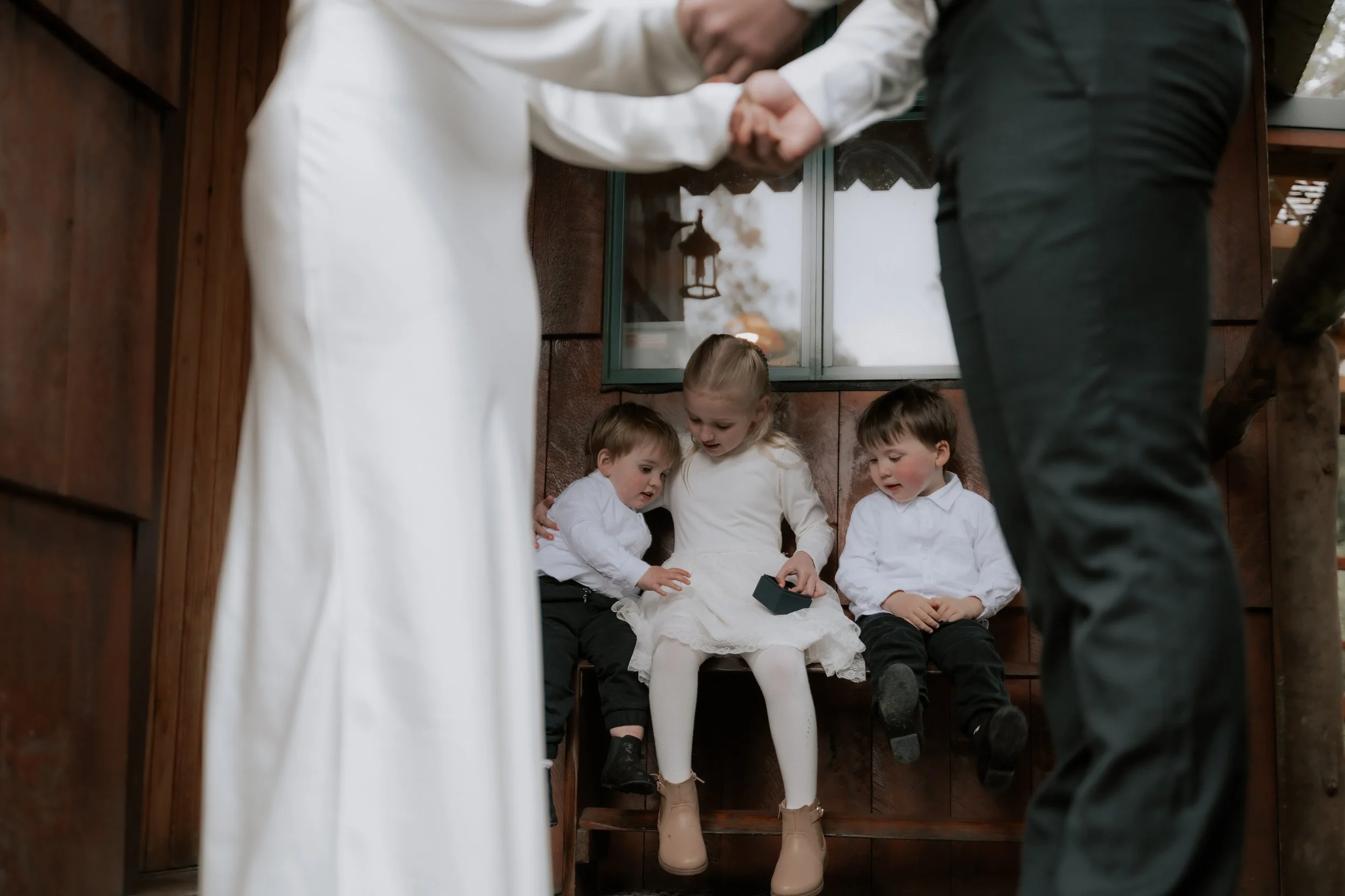 Four young children sitting in a small wooden booth with one girl in a white dress holding a small black box, two boys in white shirts next to her, and a girl with blonde hair in a white shirt, all looking at the girl with the box. Two adults are sha