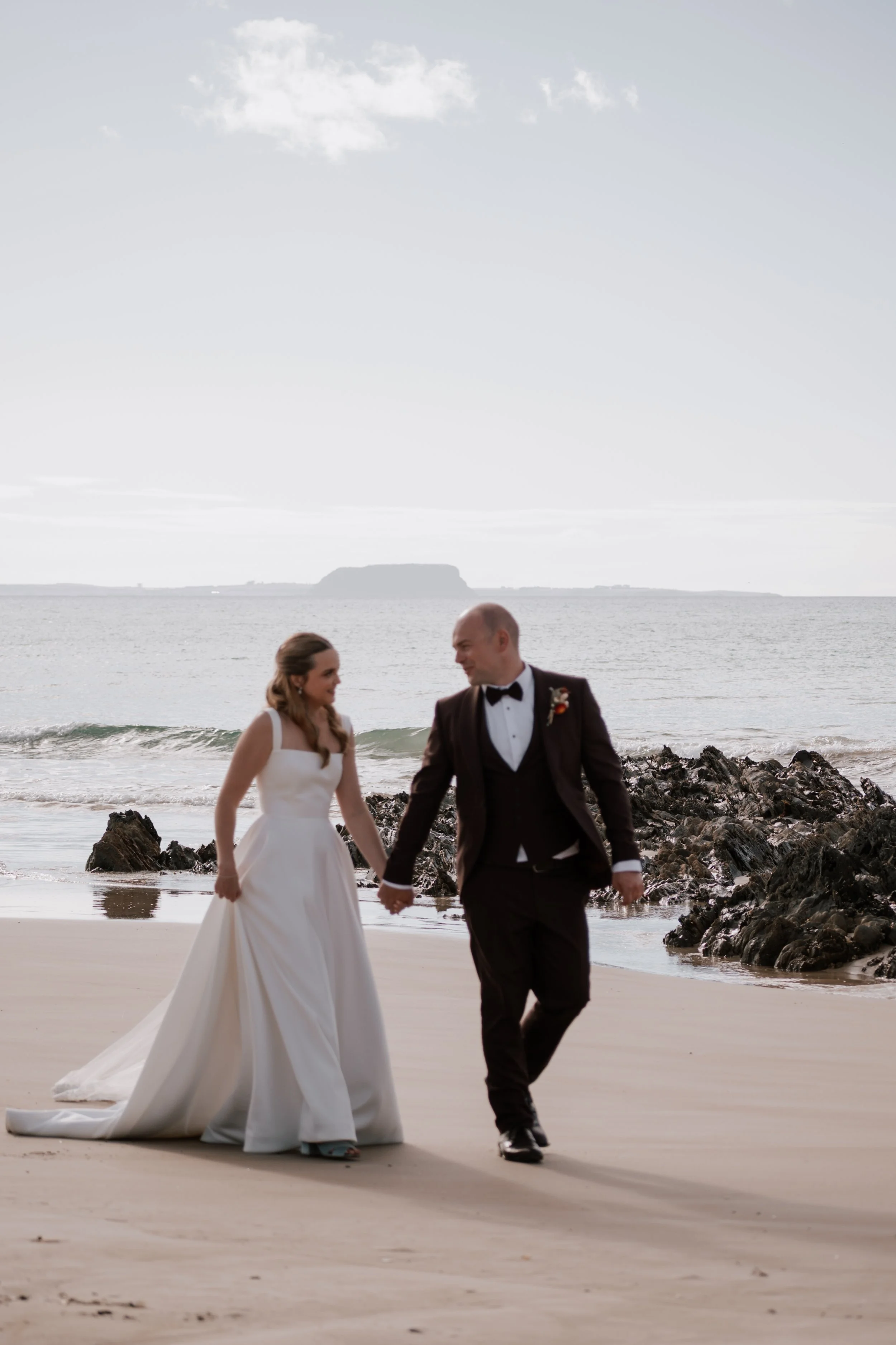 A bride and groom walking hand in hand on the beach, dressed in wedding attire, with a rocky shoreline and ocean in the background.