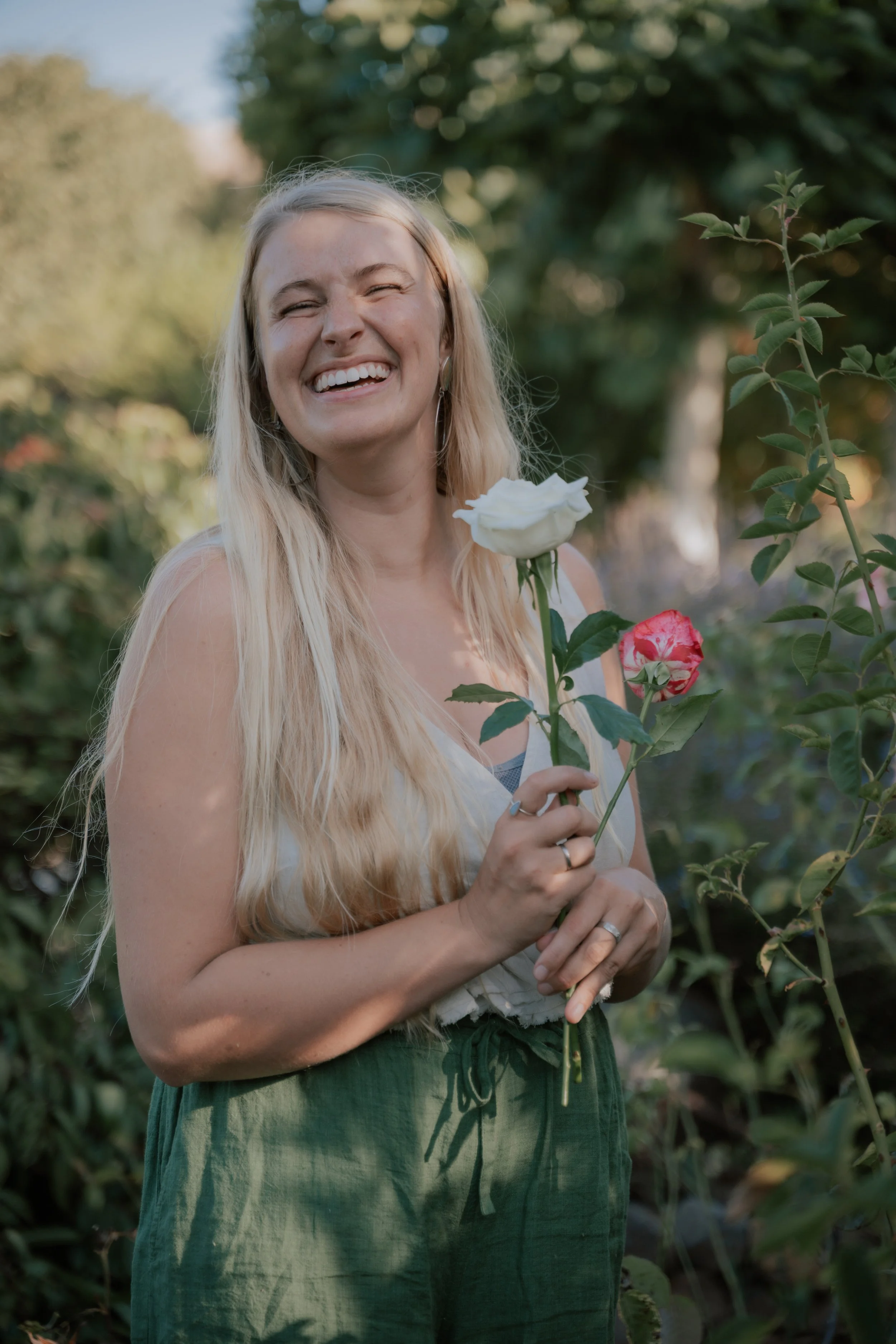 A woman with long blonde hair smiling and holding a white rose and a pink rose in a garden with green foliage.