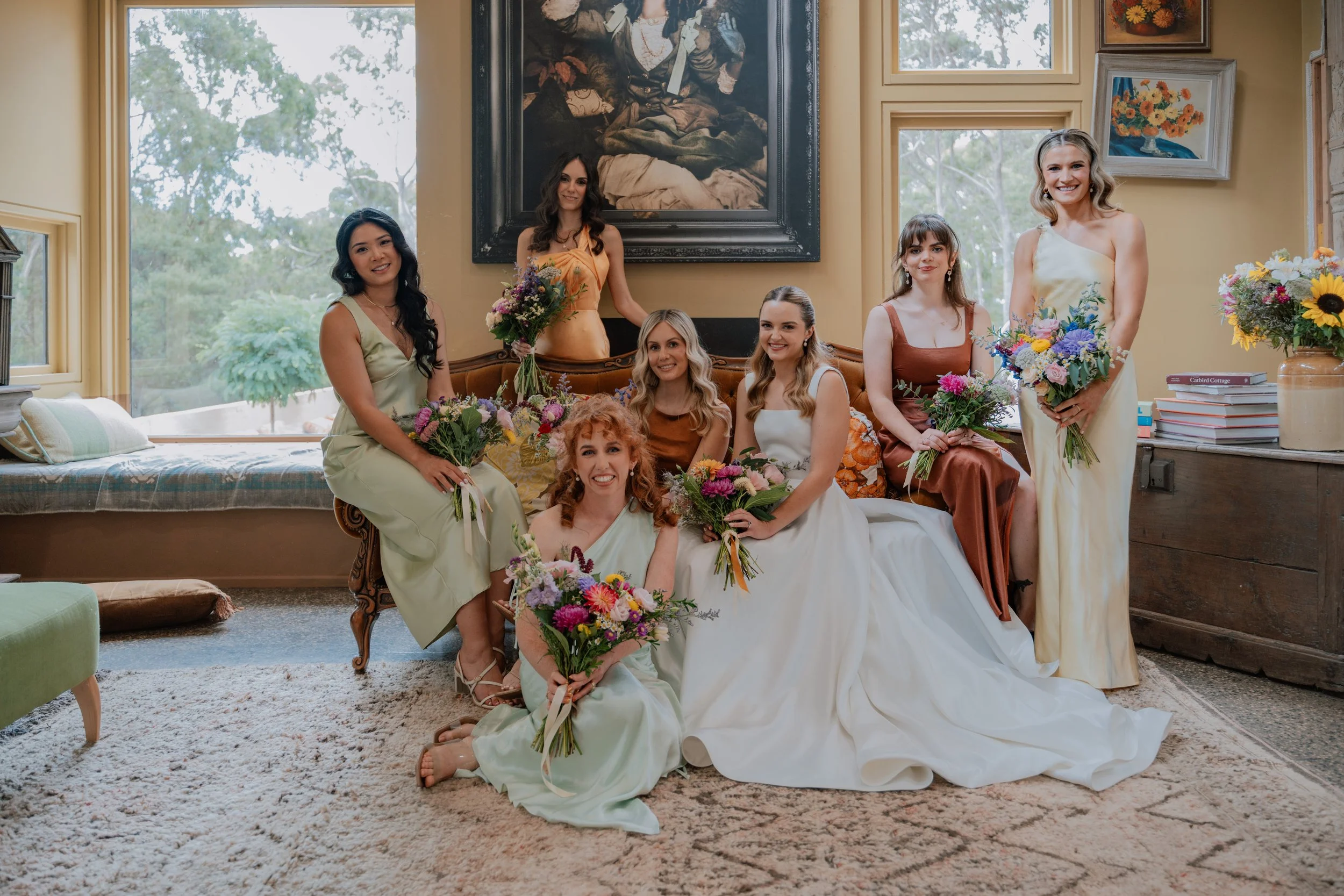 Group of seven women in colorful dresses holding bouquets, sitting and standing in a warmly lit room with large windows, botanical artwork, and a portrait hanging on the wall.