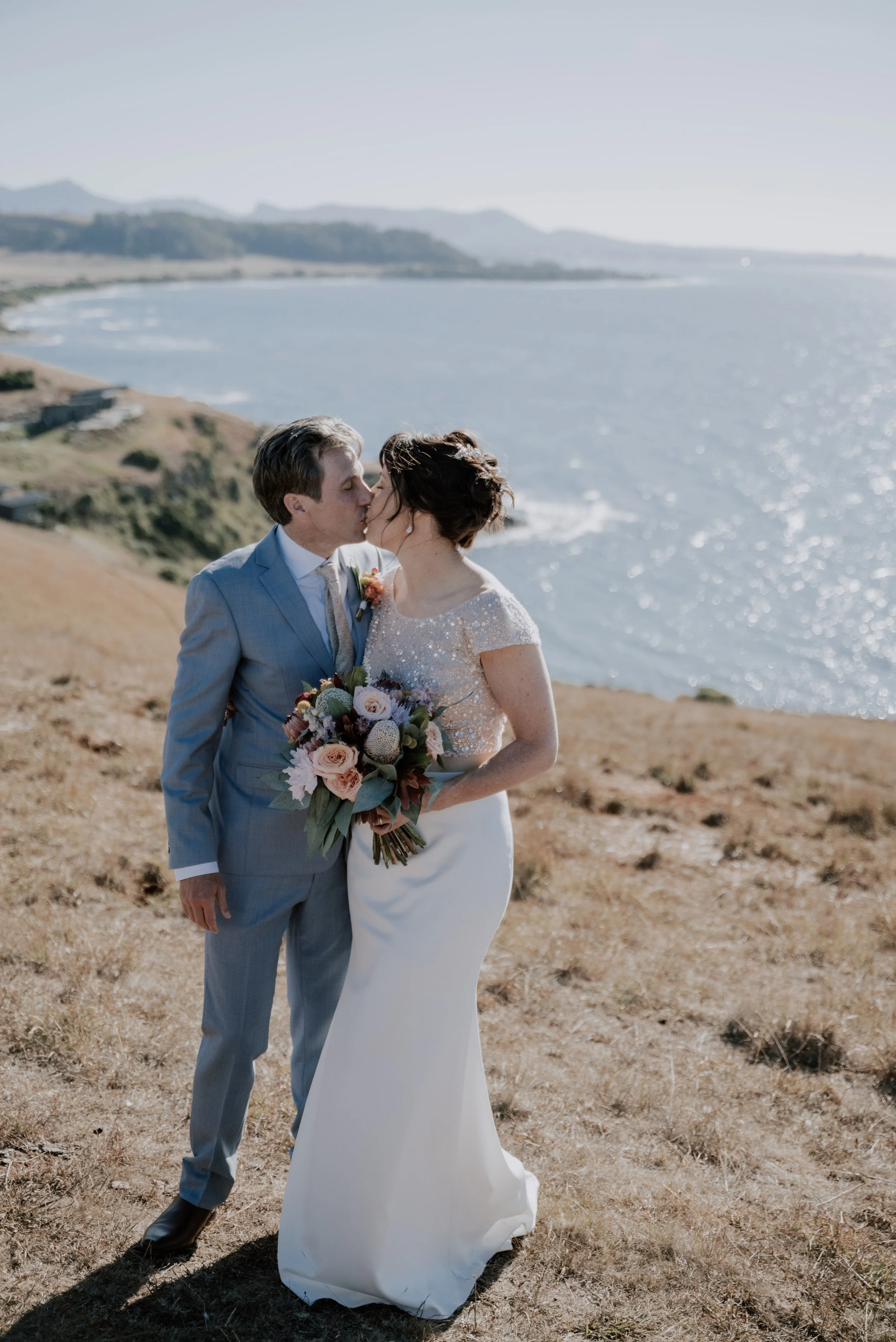 A bride and groom kiss on the hillside above the ocean with mountains in the distance