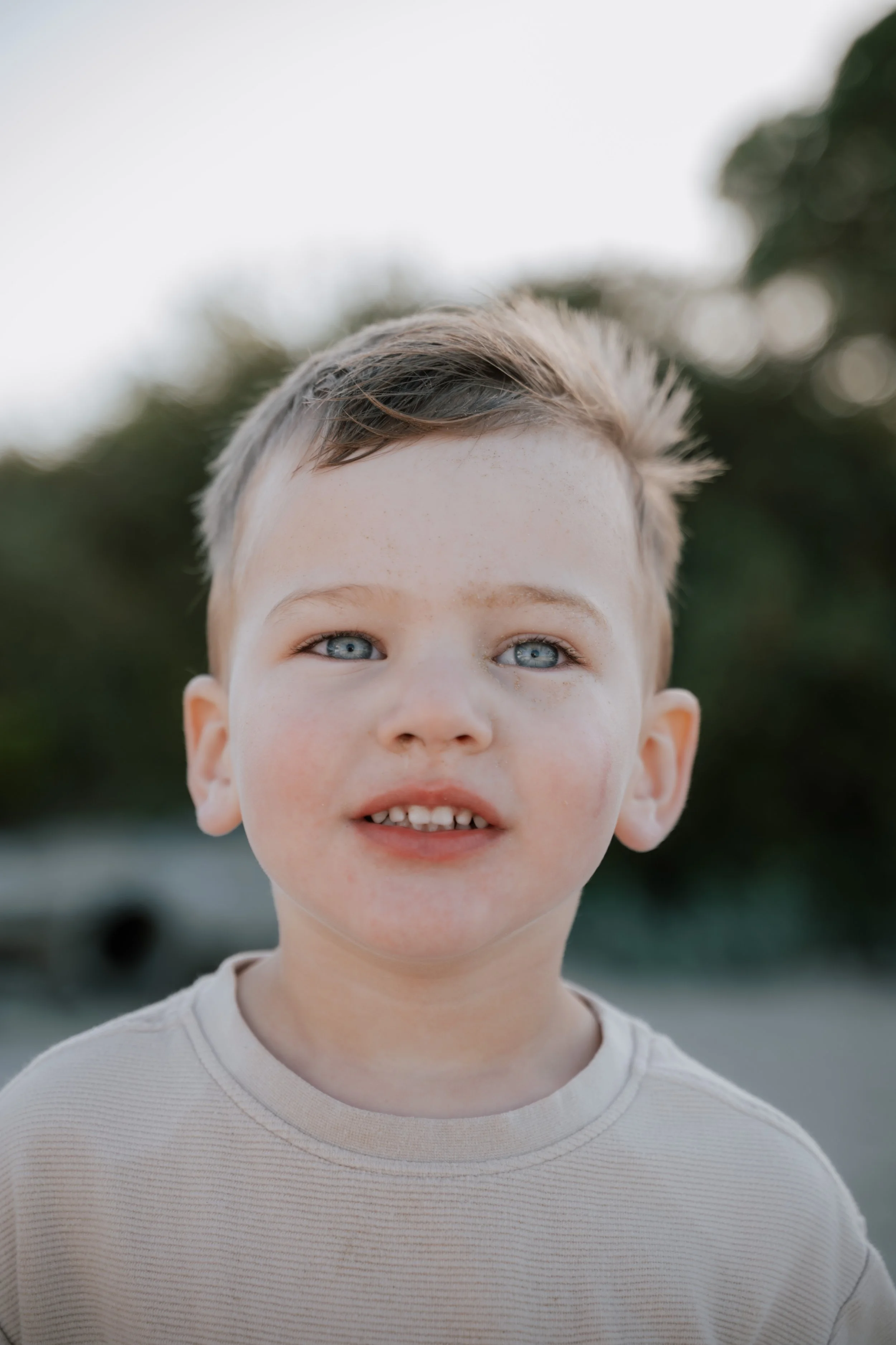 Close-up of a young boy with blue eyes, light skin, short brown hair, and a beige shirt, outdoors in a natural setting with blurred greenery in the background.