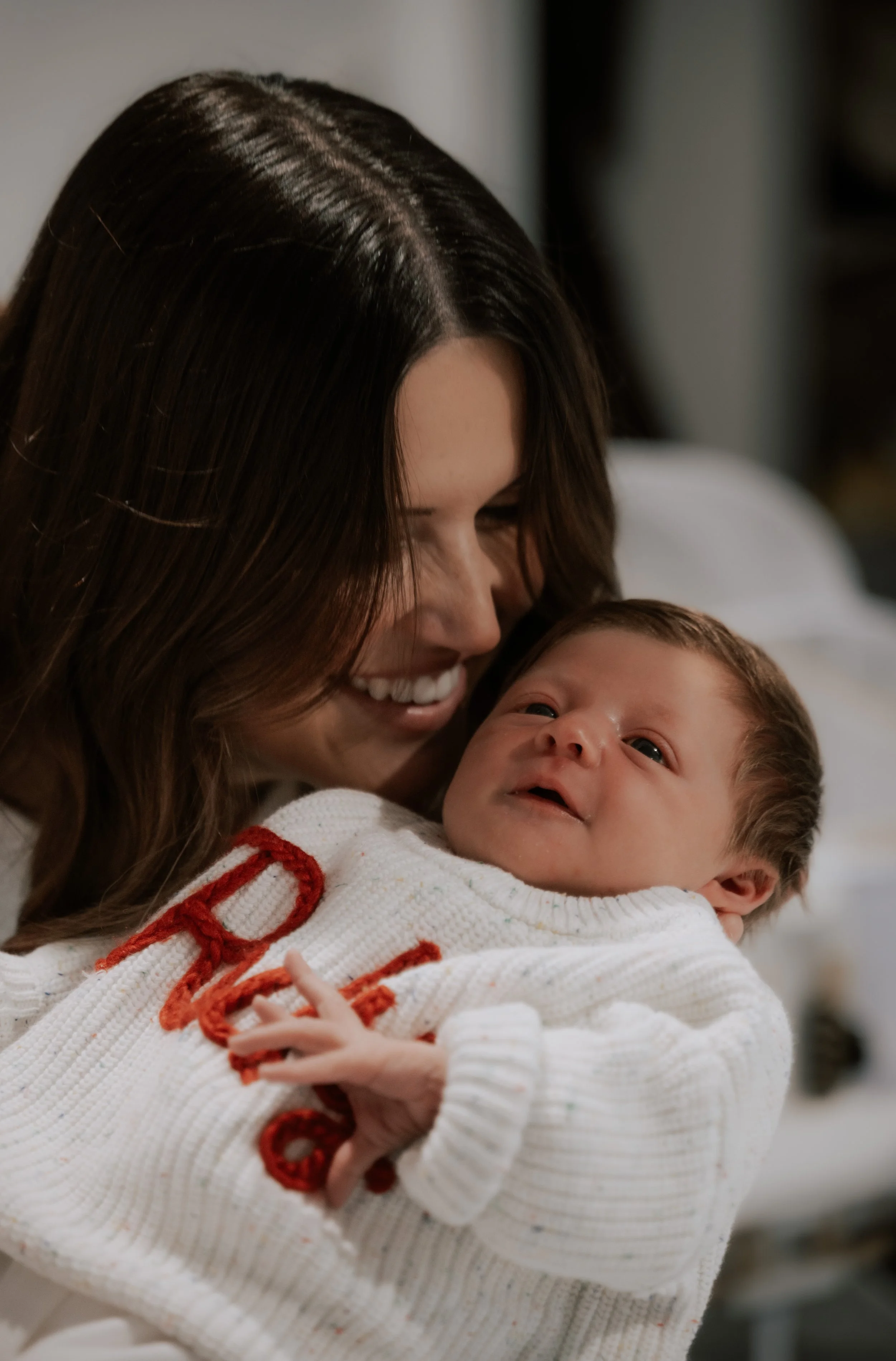A woman with long brown hair smiling and holding a baby dressed in a white sweater with red embroidery, in a warm, indoor setting.
