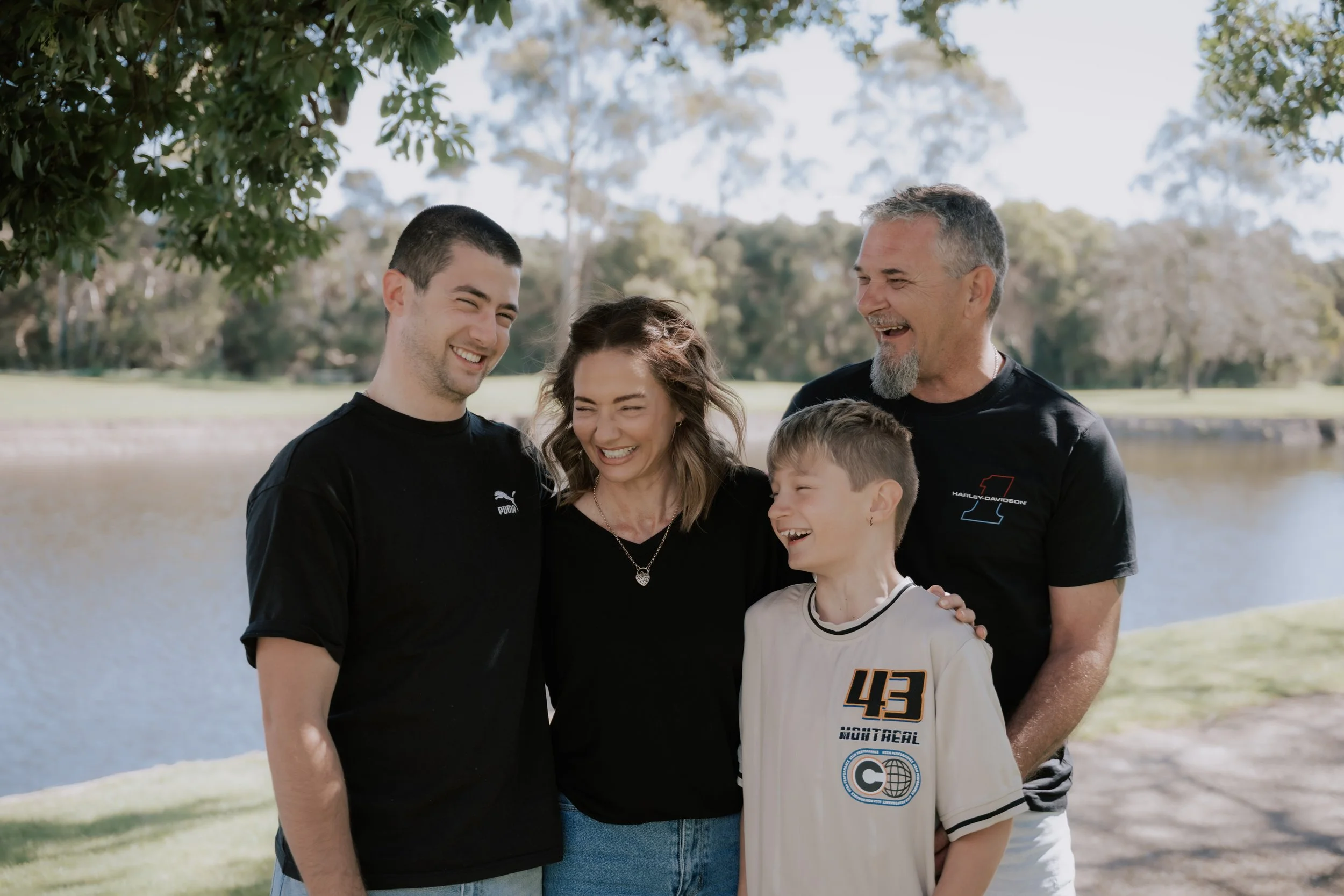 A family of five outside near a body of water, smiling and hugging each other, with trees in the background.