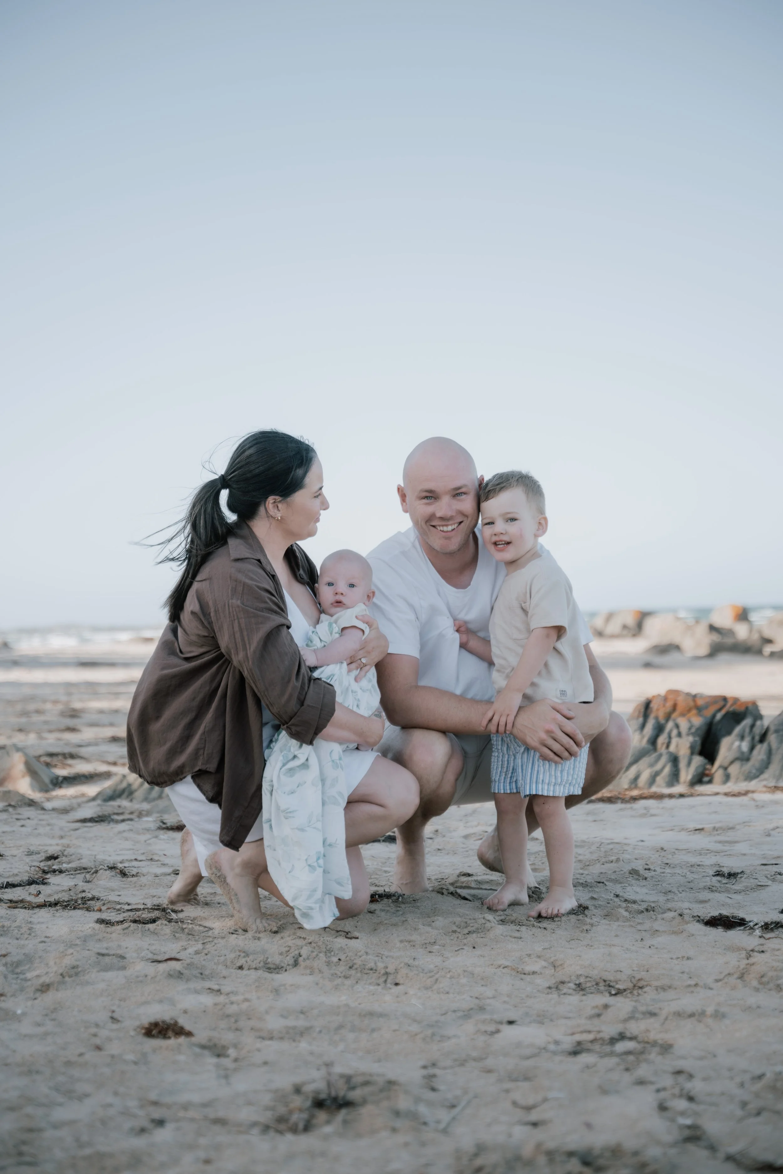Family of four at the beach, with two children and two adults, standing on the sand near rocks, smiling and enjoying the outdoors during daytime.
