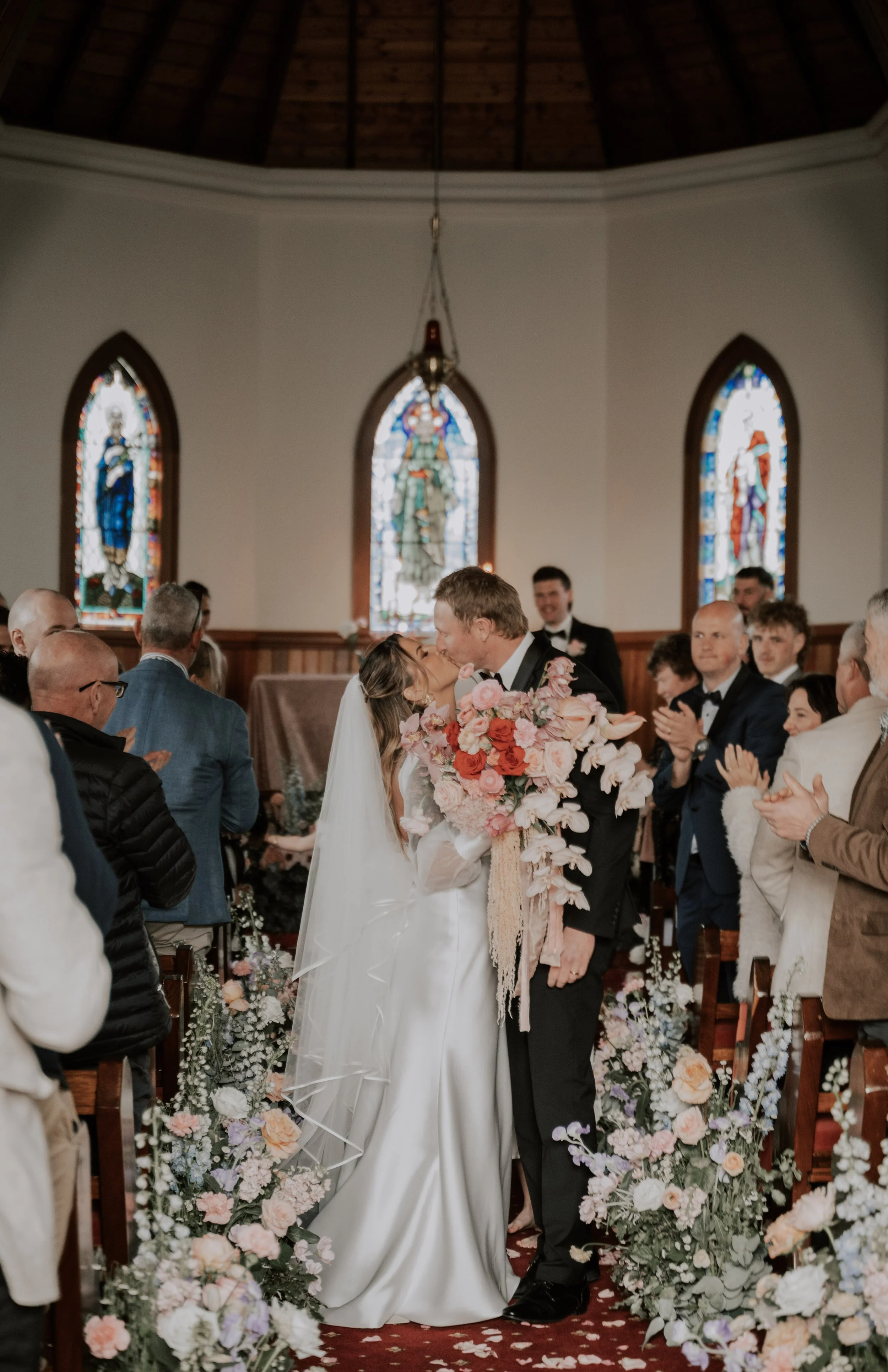 A bride and groom kiss in the aisle of an old church with stain glass windows and red carpet, surrounded by large floral components