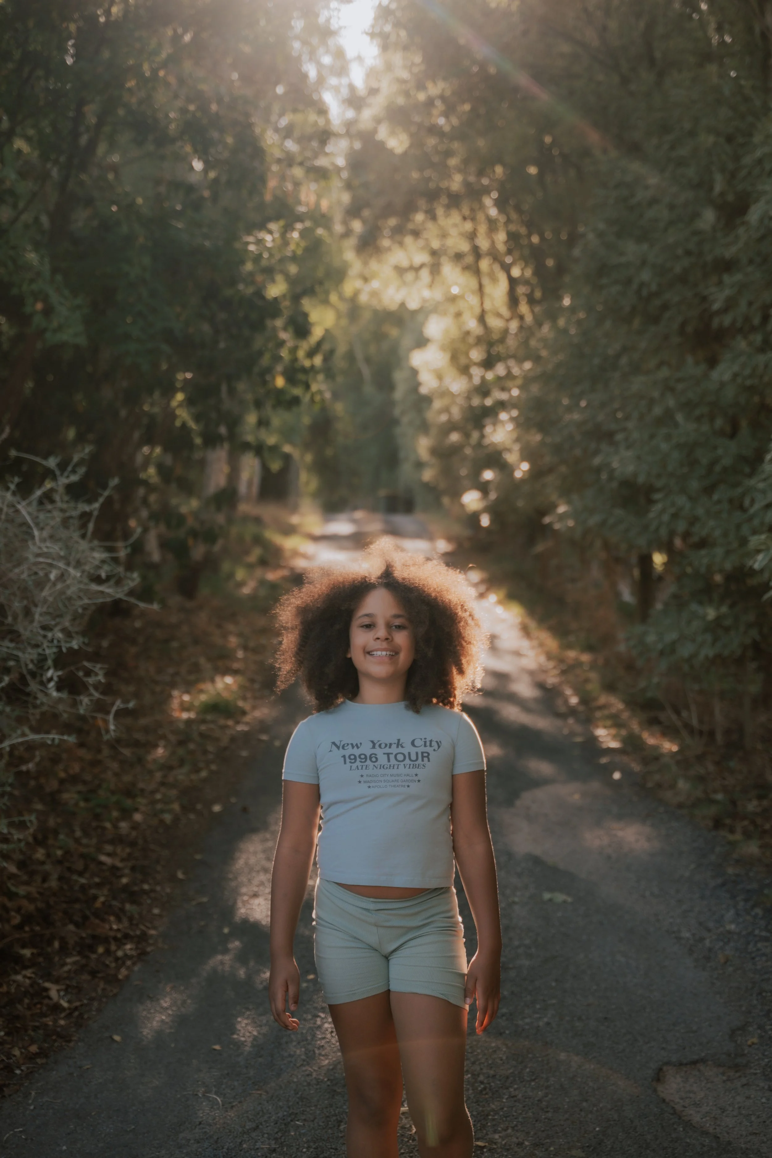 A young girl with curly hair smiling, walking on a dirt path in a wooded area during sunset, wearing a white t-shirt with text and shorts.