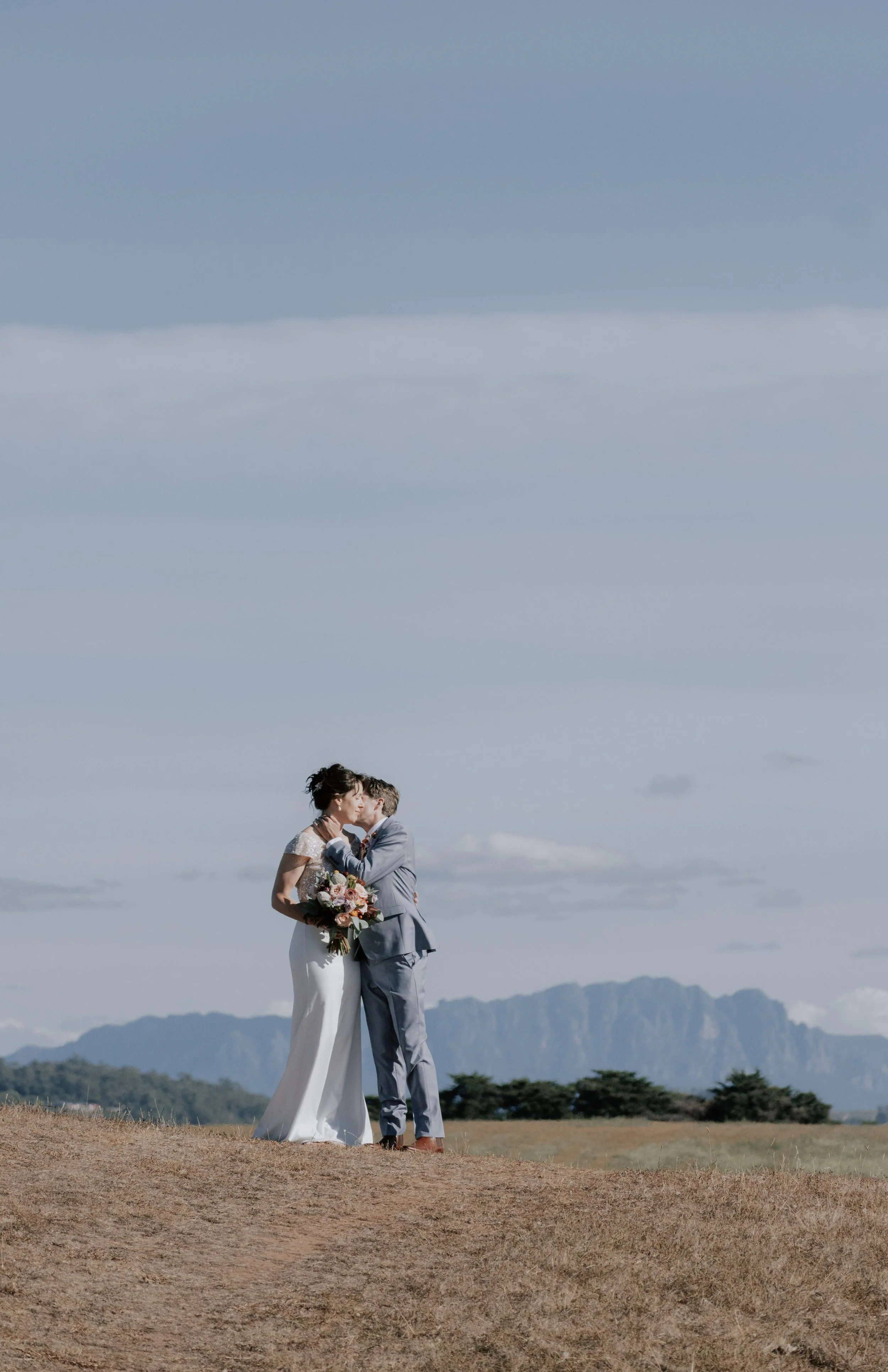 A just married bride and groom stand in the distance in a field with Mt Roland behind them and a blue sky