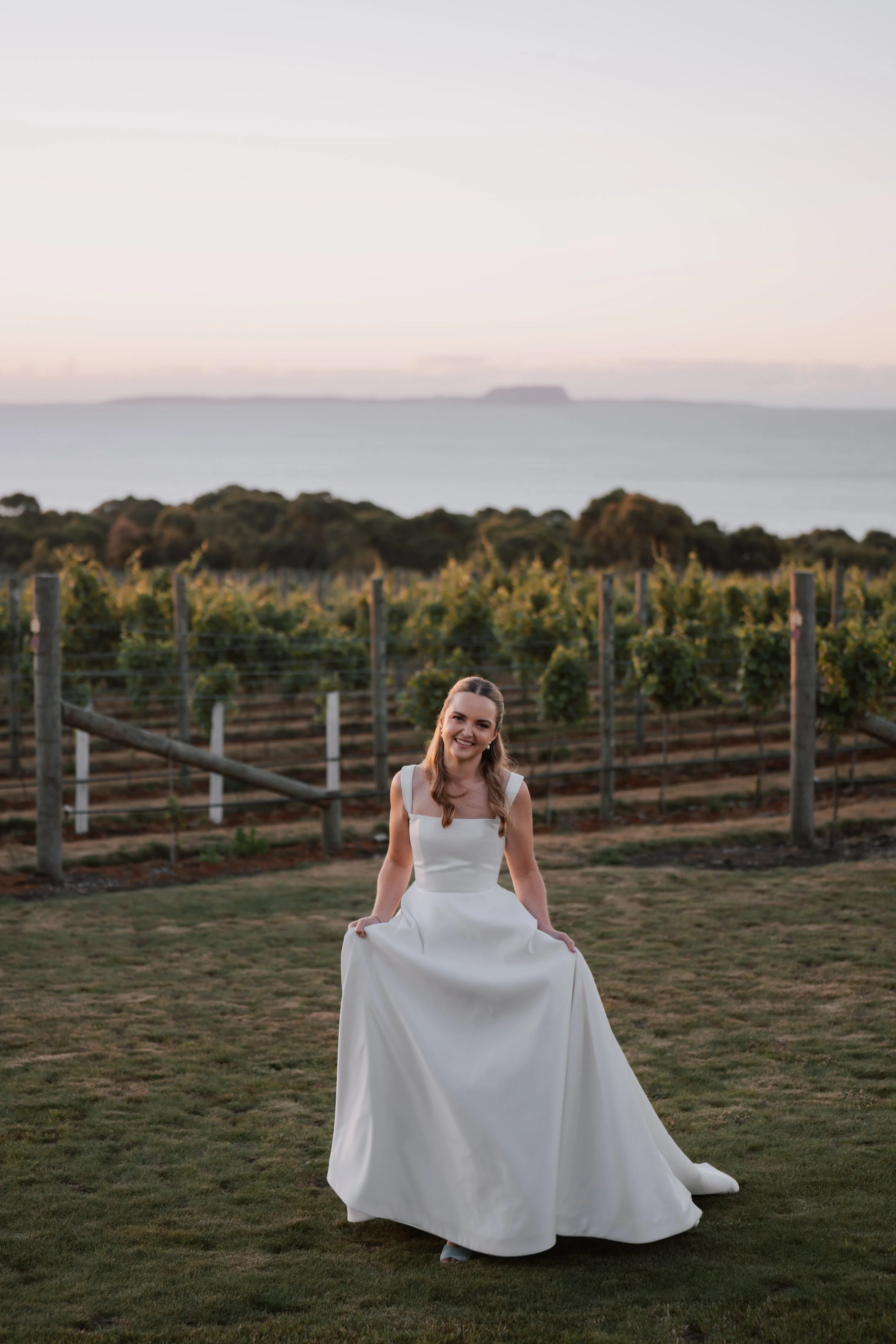A young woman in a white wedding dress sitting on the grass in a vineyard during sunset.