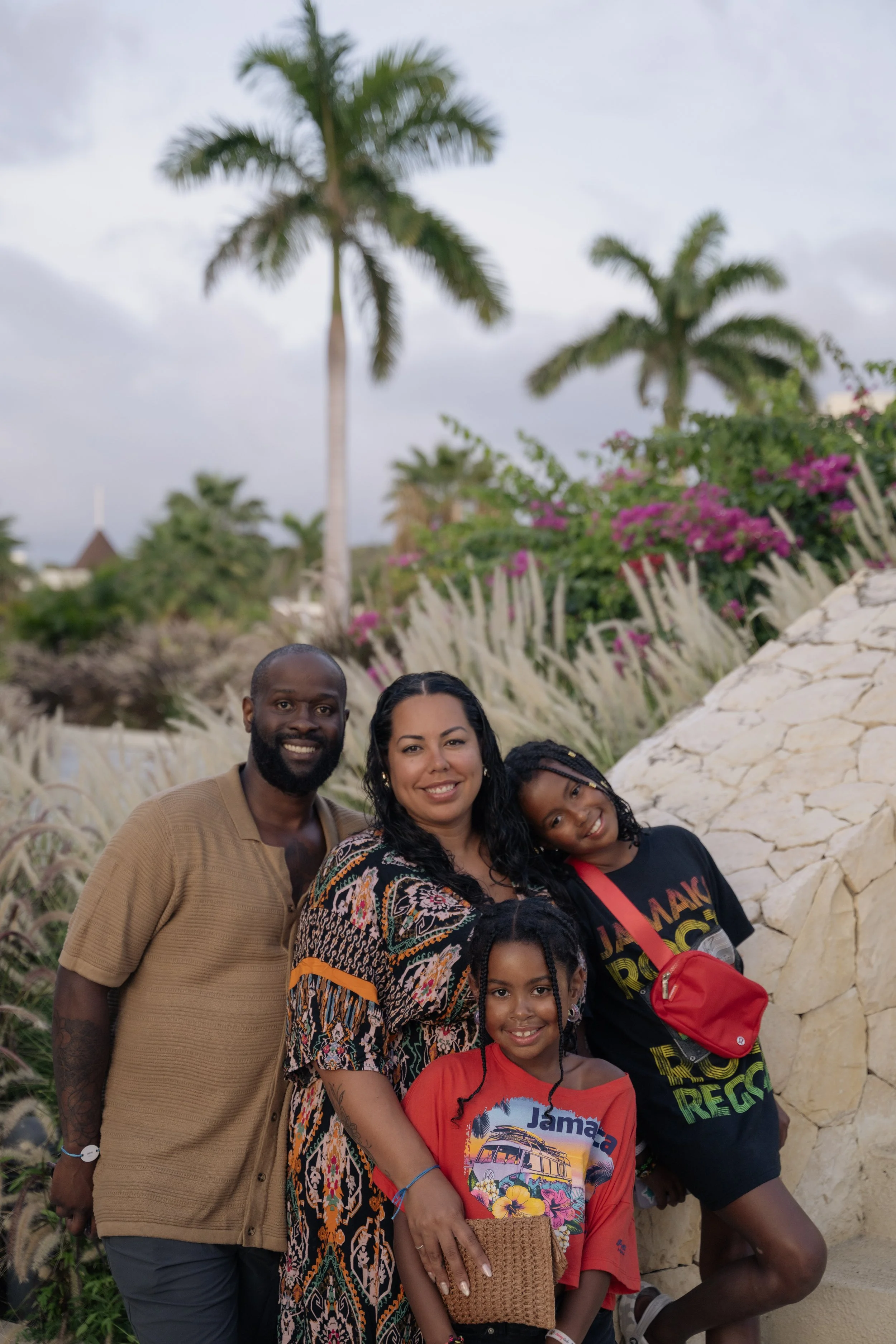 A young black family smiles at the camera, two girls and their mother and father, in front of palm trees and colourful plants