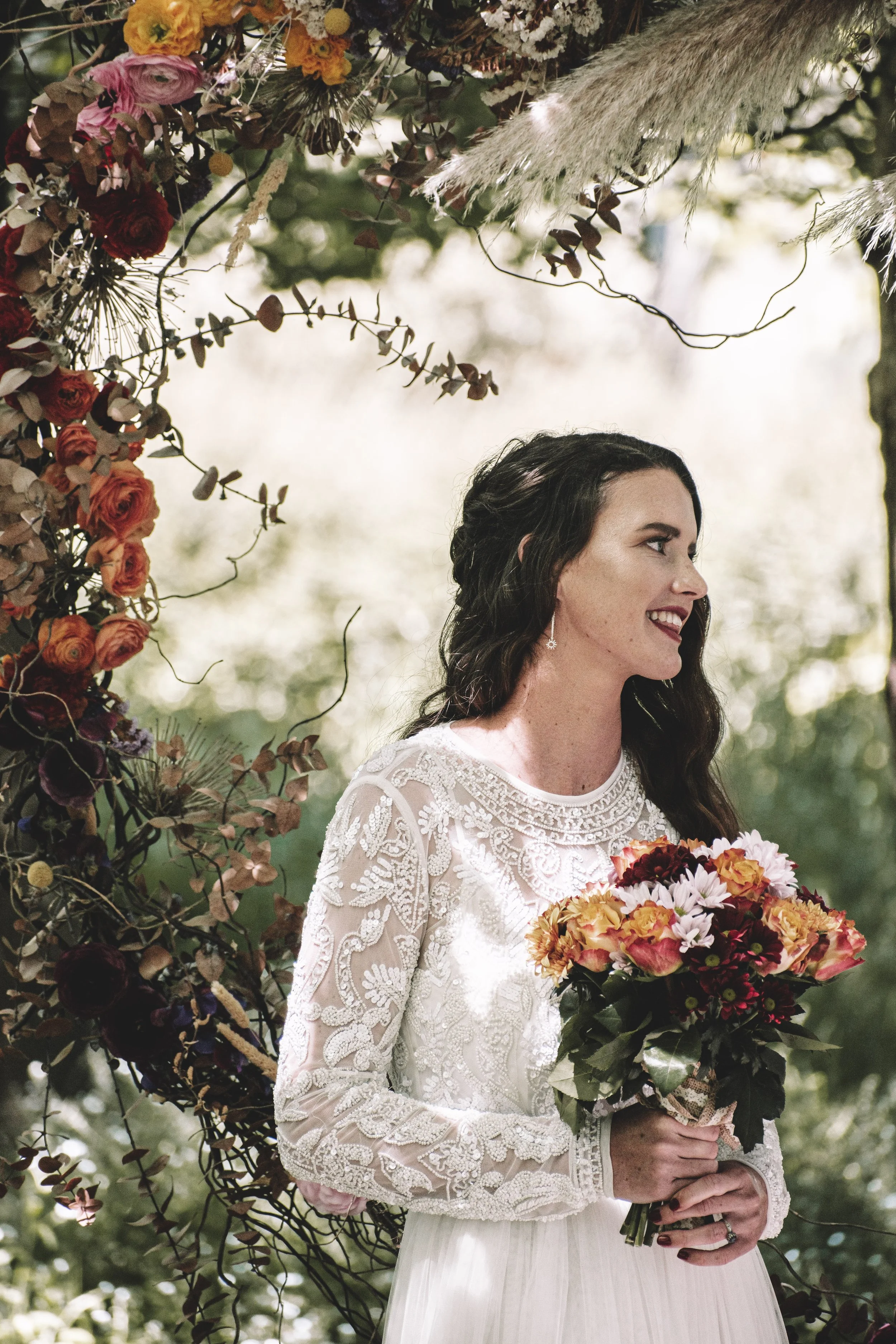 A brunette bride, in a long sleeved lace dress smiles to the side while standing in front of floral wreath in a forest