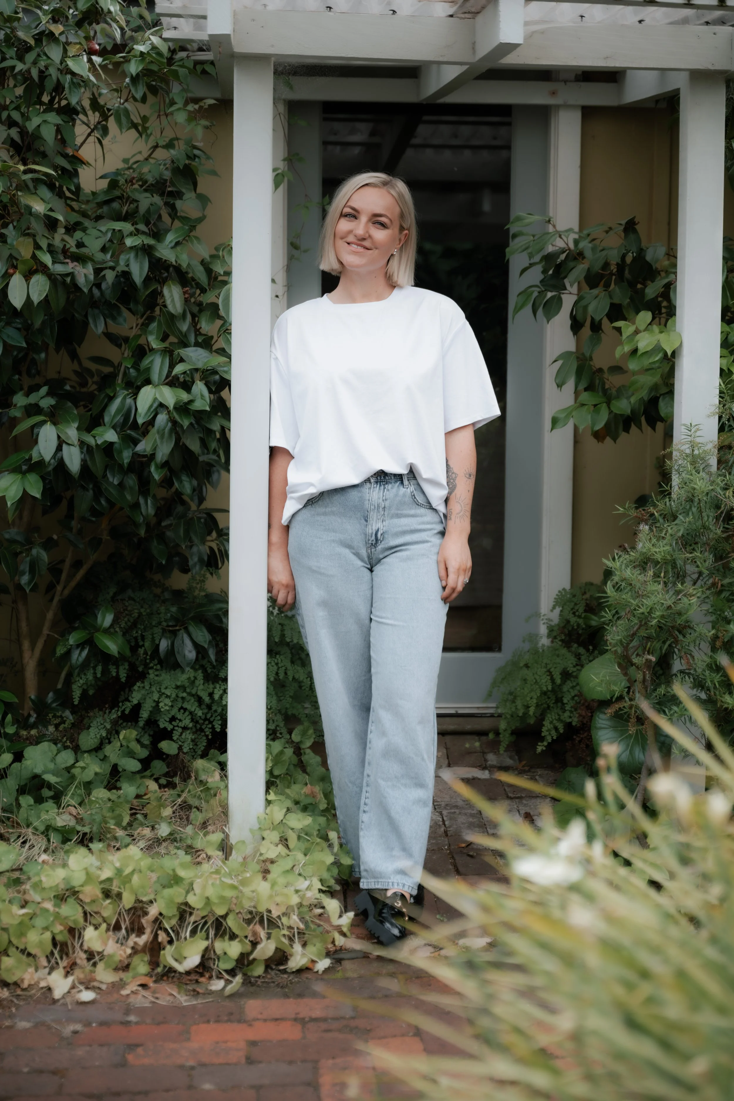 A woman with blonde hair wearing a white t-shirt, light blue jeans, and black shoes standing outdoors in front of a wooden pergola with greenery around.