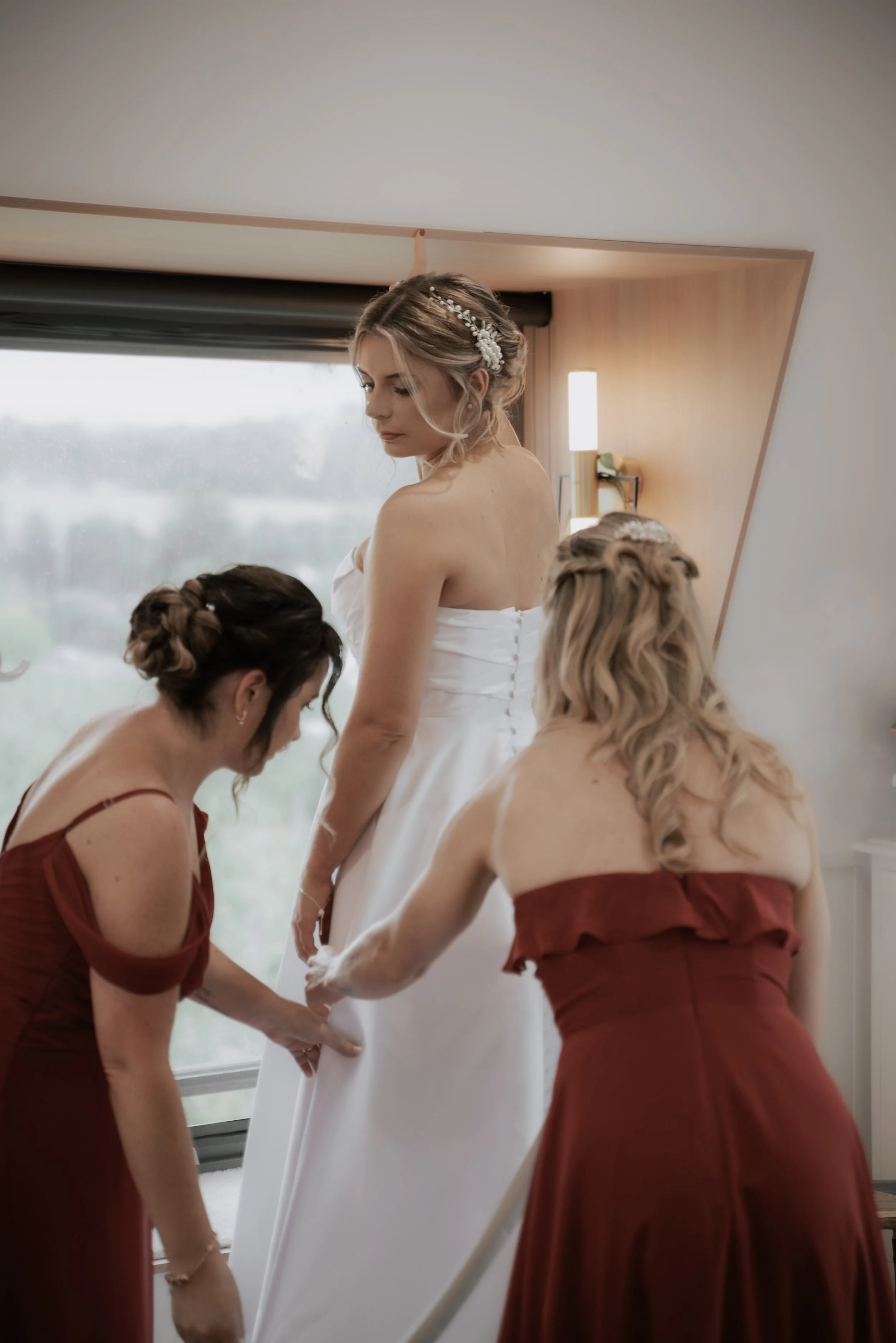 A bride being assisted by her bridesmaids in red dresses, indoors in warm lighting