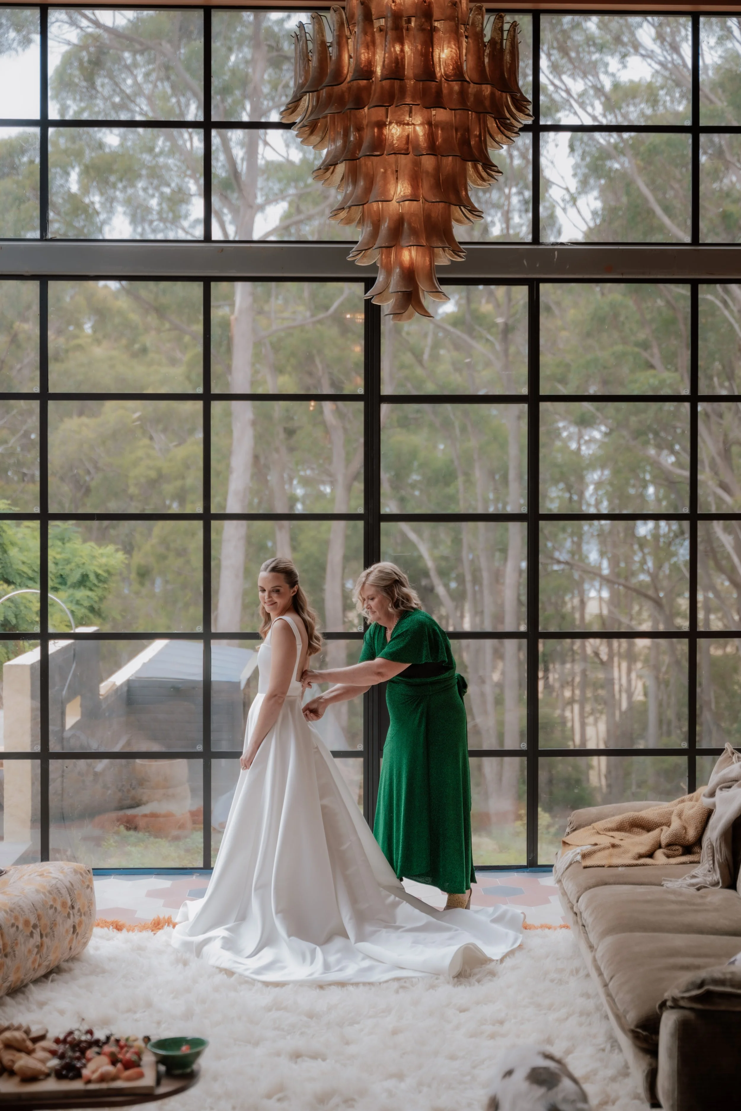 A woman in a white wedding dress is standing on a fluffy white rug inside a room with large grid-style windows. An older woman in a green dress is helping her with her dress. There is a large copper-colored chandelier hanging from the ceiling, and lu