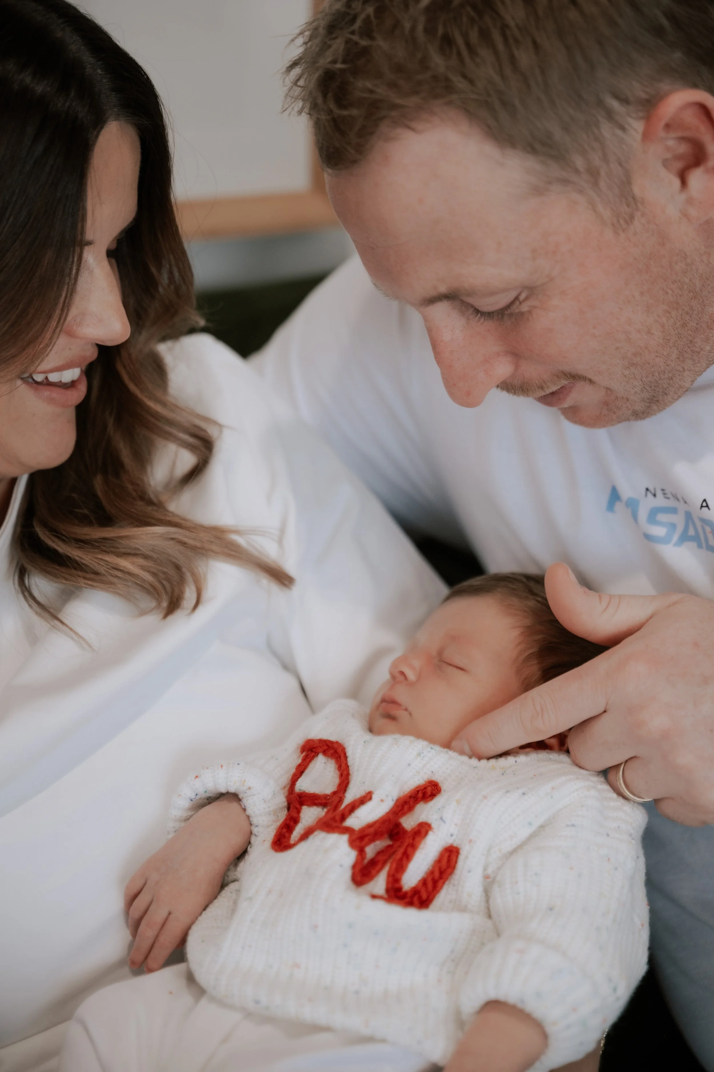 A woman and a man are holding a sleeping baby, smiling and looking at the baby in a cozy setting.