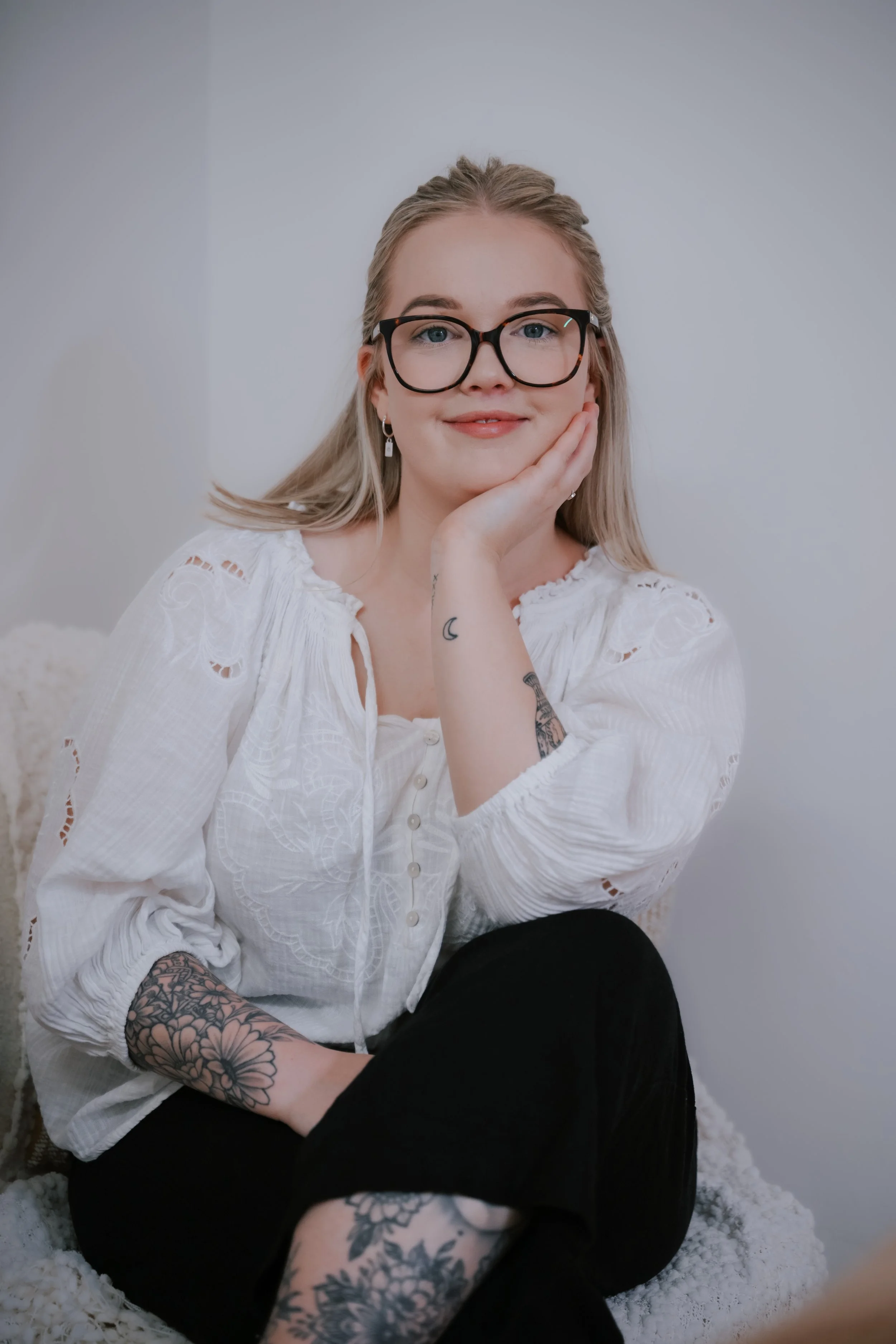 A woman with blonde hair, glasses, and tattoos sitting on a fluffy rug against a white wall, resting her face on her hand and smiling.