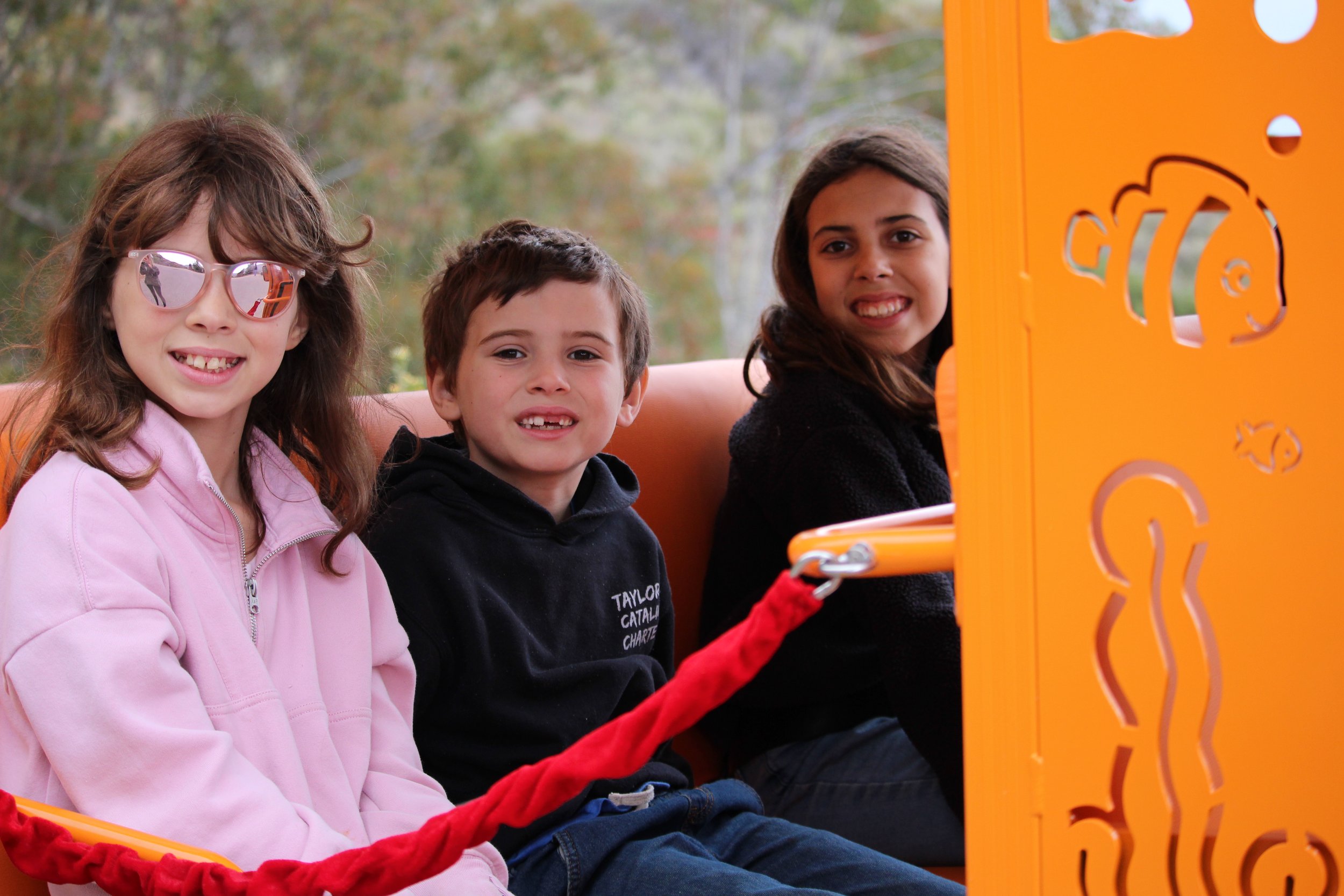 Three children sitting on a bus with trees visible in the background, beside an orange divider with bear and various decorative cutouts.