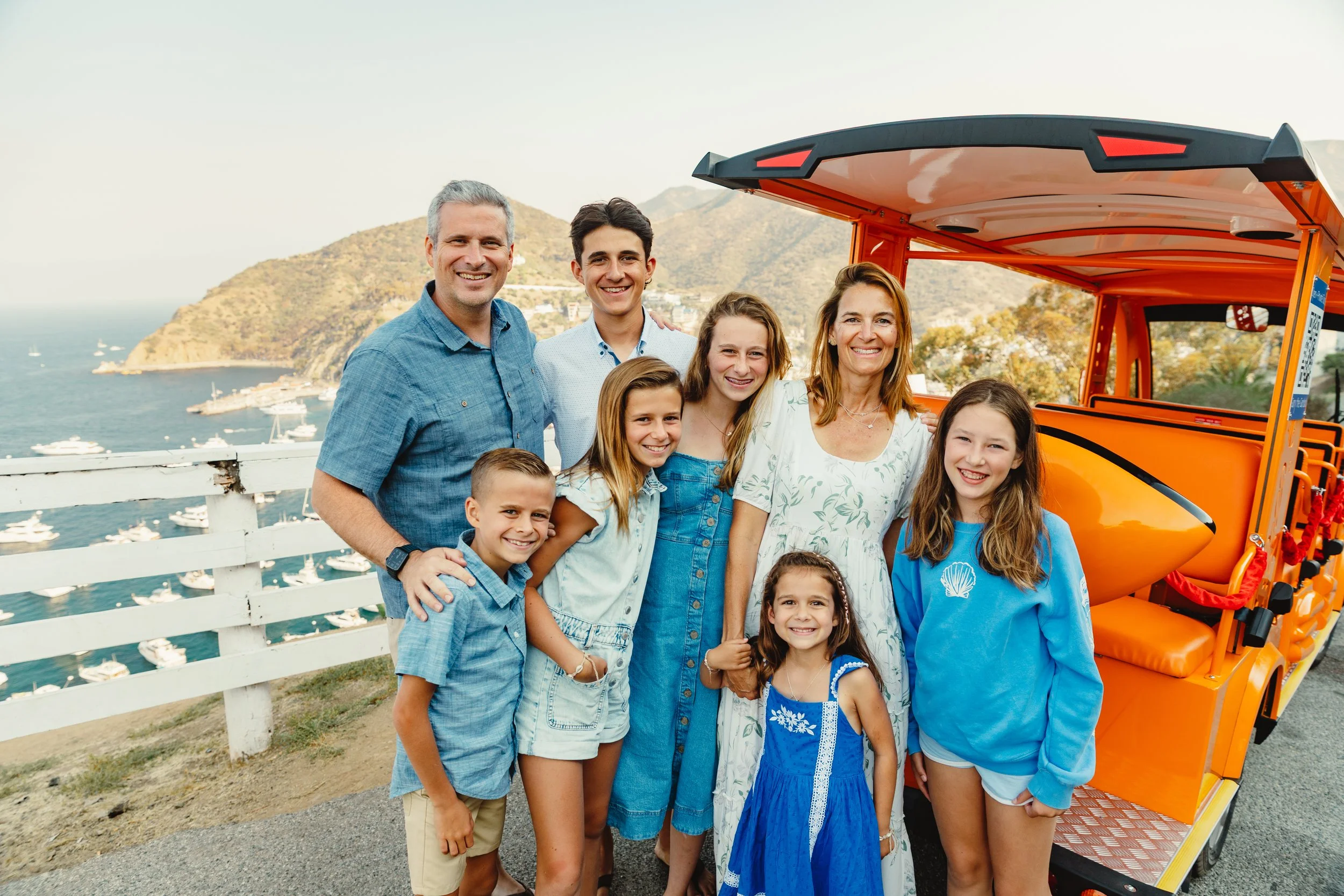 Family of nine smiling outdoors on a sightseeing ride near a harbor with boats, mountainous landscape in the background, standing next to an orange tram or trolley.