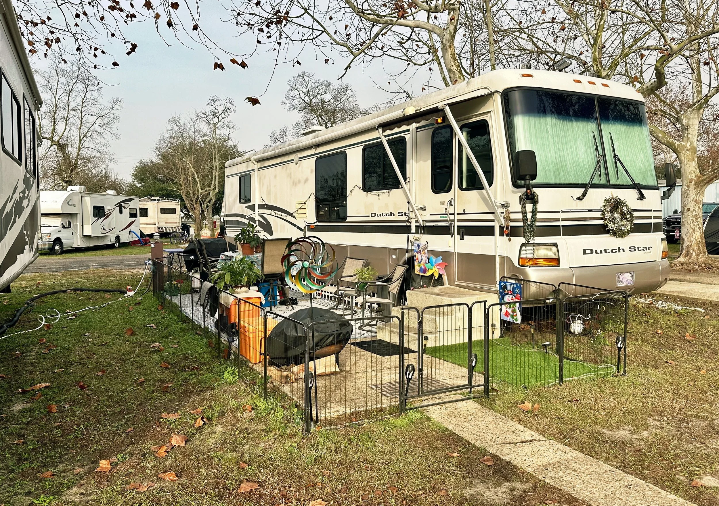A recreational vehicle (RV) parked at an RV park with various outdoor decorations and furniture, including a small fenced patio area, plants, wind spinners, a small artificial grass patch, and holiday decorations. There are other RVs visible in the background, along with leafless trees and an overcast sky.