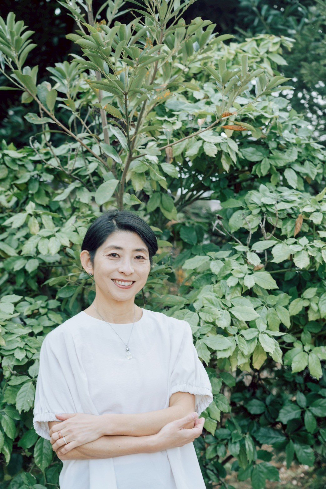 A woman with short black hair, wearing a white dress and a necklace, smiling and standing in front of lush green foliage.