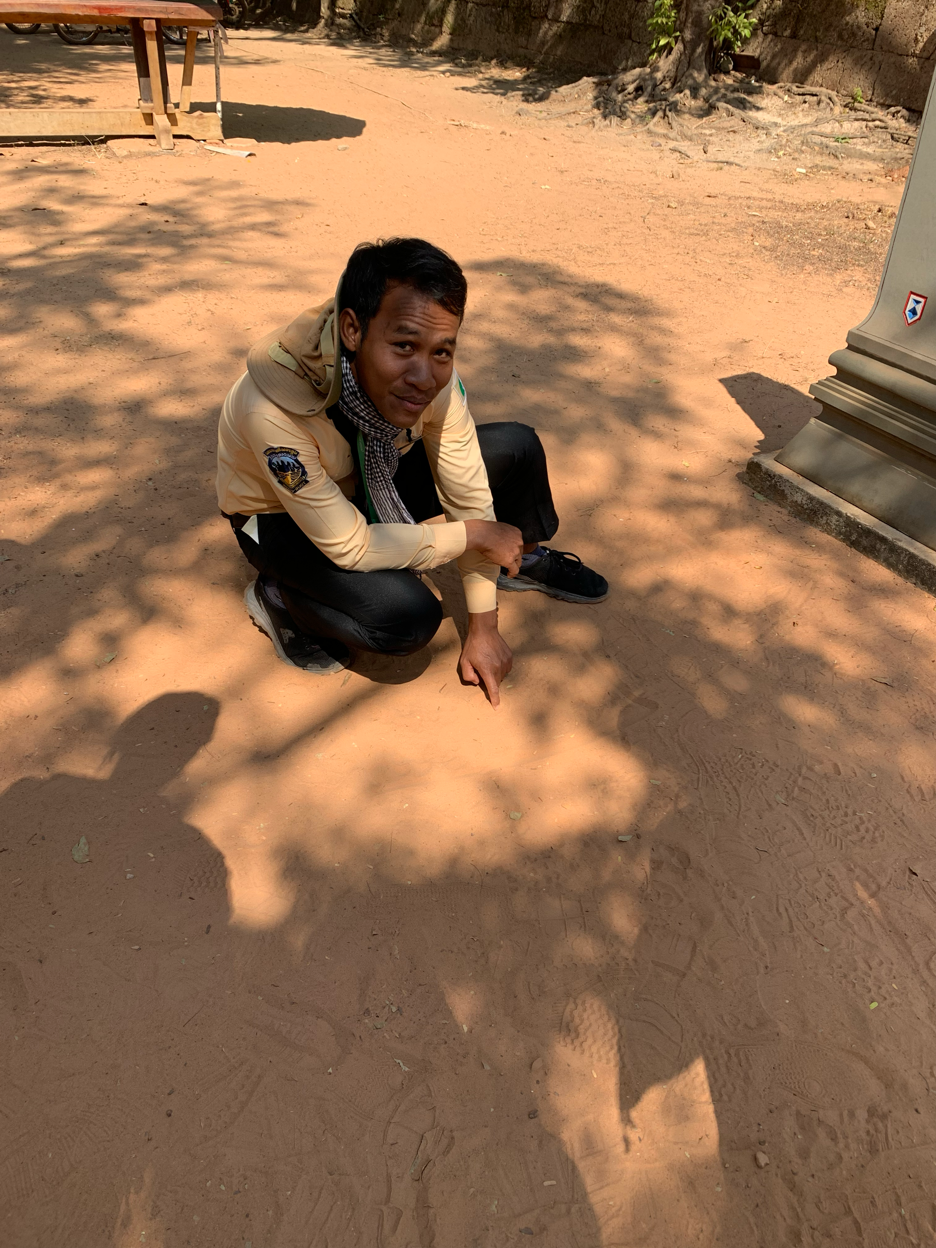 A man kneeling on the ground in a sandy outdoor area, pointing at the ground with his right finger, with shadows cast on the ground.