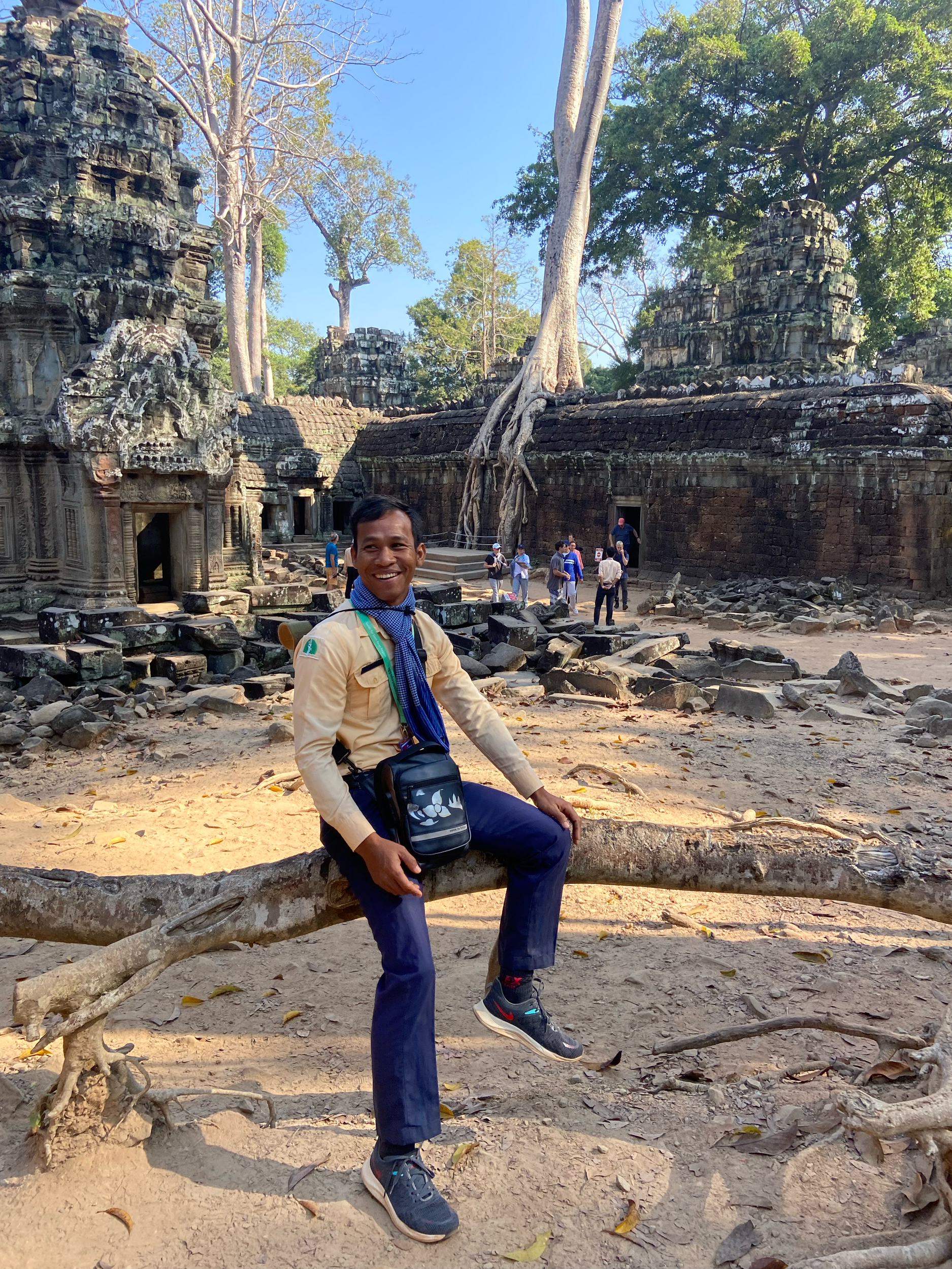 A smiling man sitting on a tree branch in front of ancient stone temple ruins with large trees overhead and a group of tourists exploring in the background.