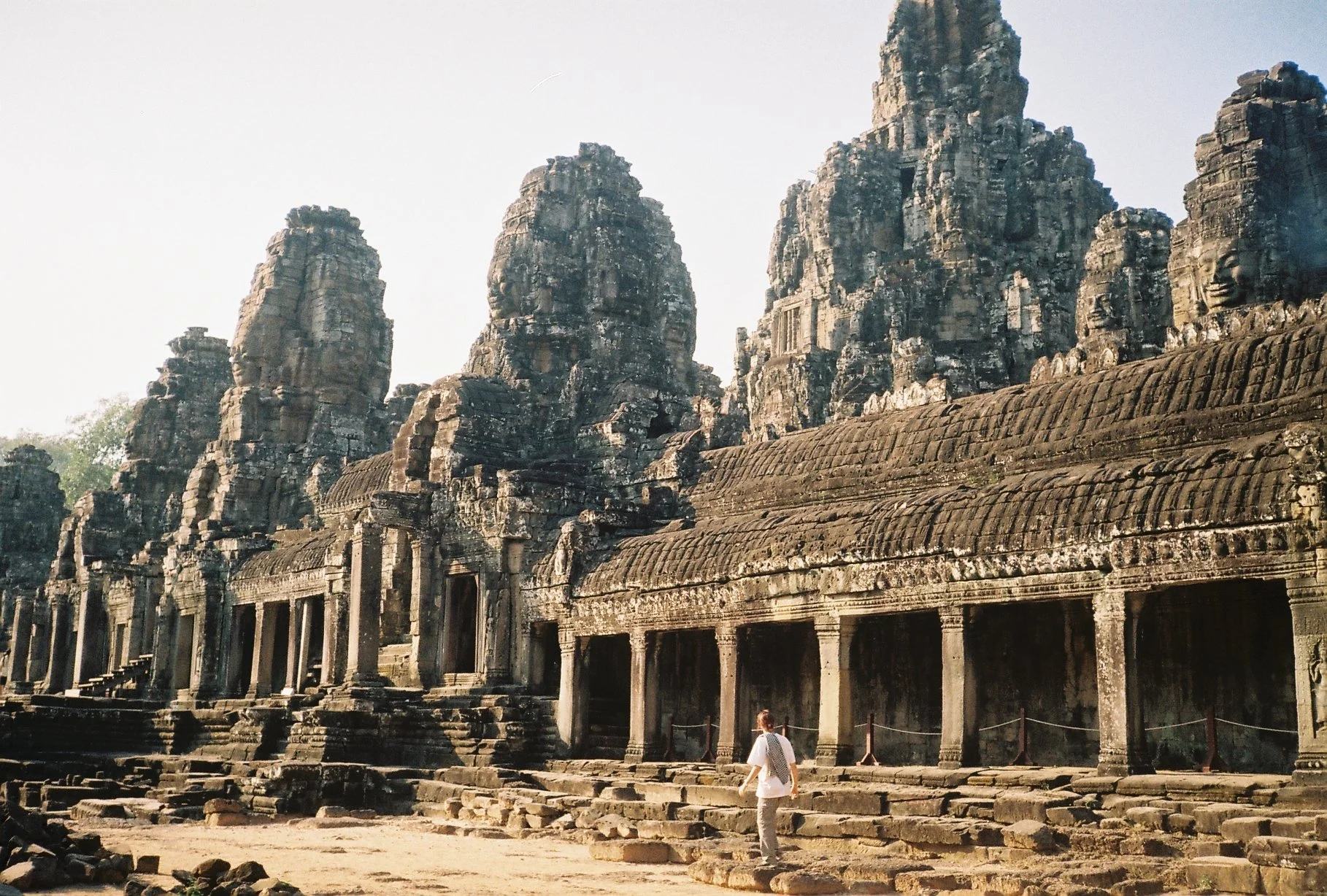 Ancient stone temple ruins with carved faces on towers and a person walking in front, surrounded by steps and columns.