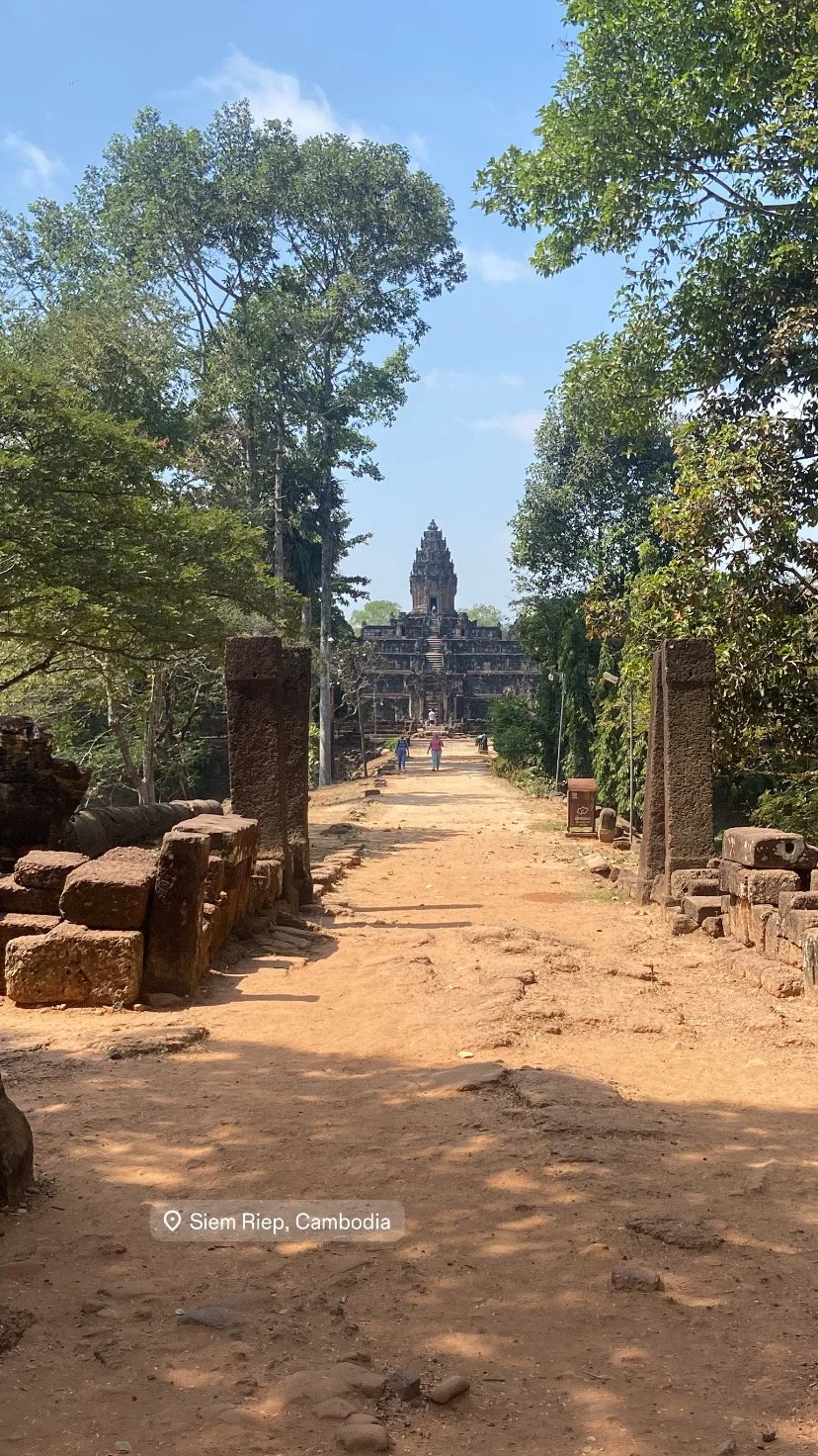 Dirt pathway leading to ancient stone temple in Siem Reap, Cambodia, bordered by trees and ruins.