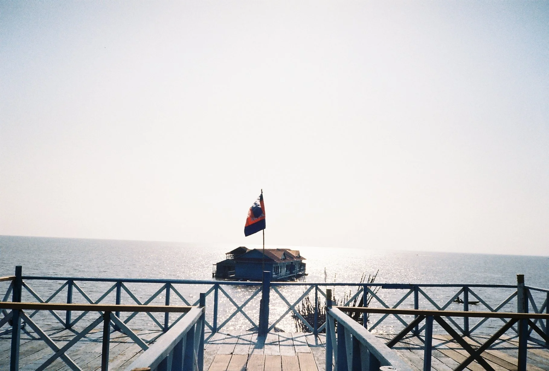 A wooden deck with white railings overlooking a shimmering sea. There is a house on stilts in the water with a flag on a pole in front of it, and the sky is clear with bright sunlight.