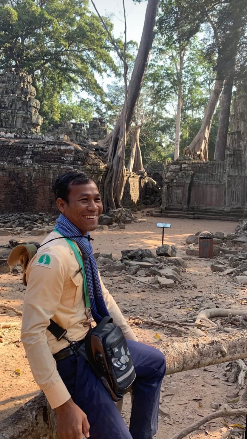 Smiling man with camera bag sitting on a log in an ancient stone temple with large trees and roots in the background.