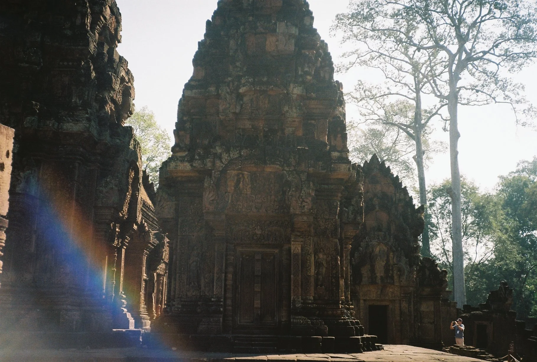 Ancient stone temple with intricate carvings, surrounded by trees and sunlight shining through, with a person taking a photo near the temple.