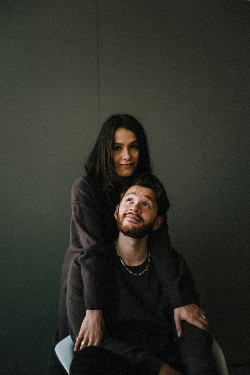 A woman with shoulder-length dark hair stands behind a seated man with dark hair and a beard, both wearing dark clothing, against a dark background.